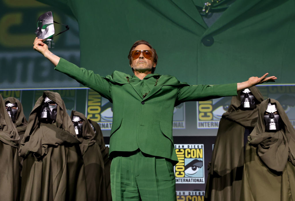 Robert Downey Jr. on stage at Comic-Con International wearing a stylish suit, holding a mask, and surrounded by individuals in cloaks and masks