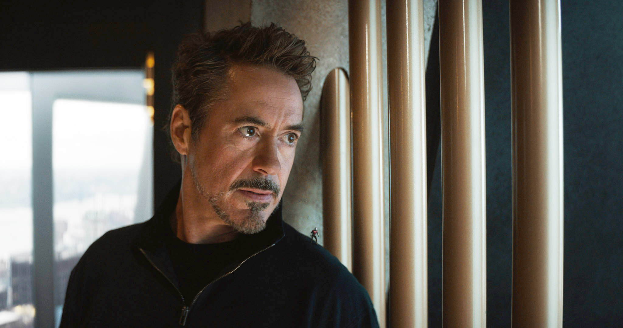 Robert Downey Jr. stands in front of cylindrical structures, looking to the left with a serious expression. He is wearing a dark jacket