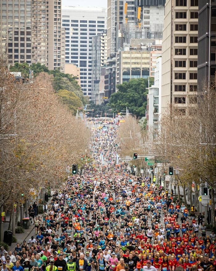 A large crowd of people running a marathon on a city street lined with buildings. Traffic lights and naked trees are visible along the route
