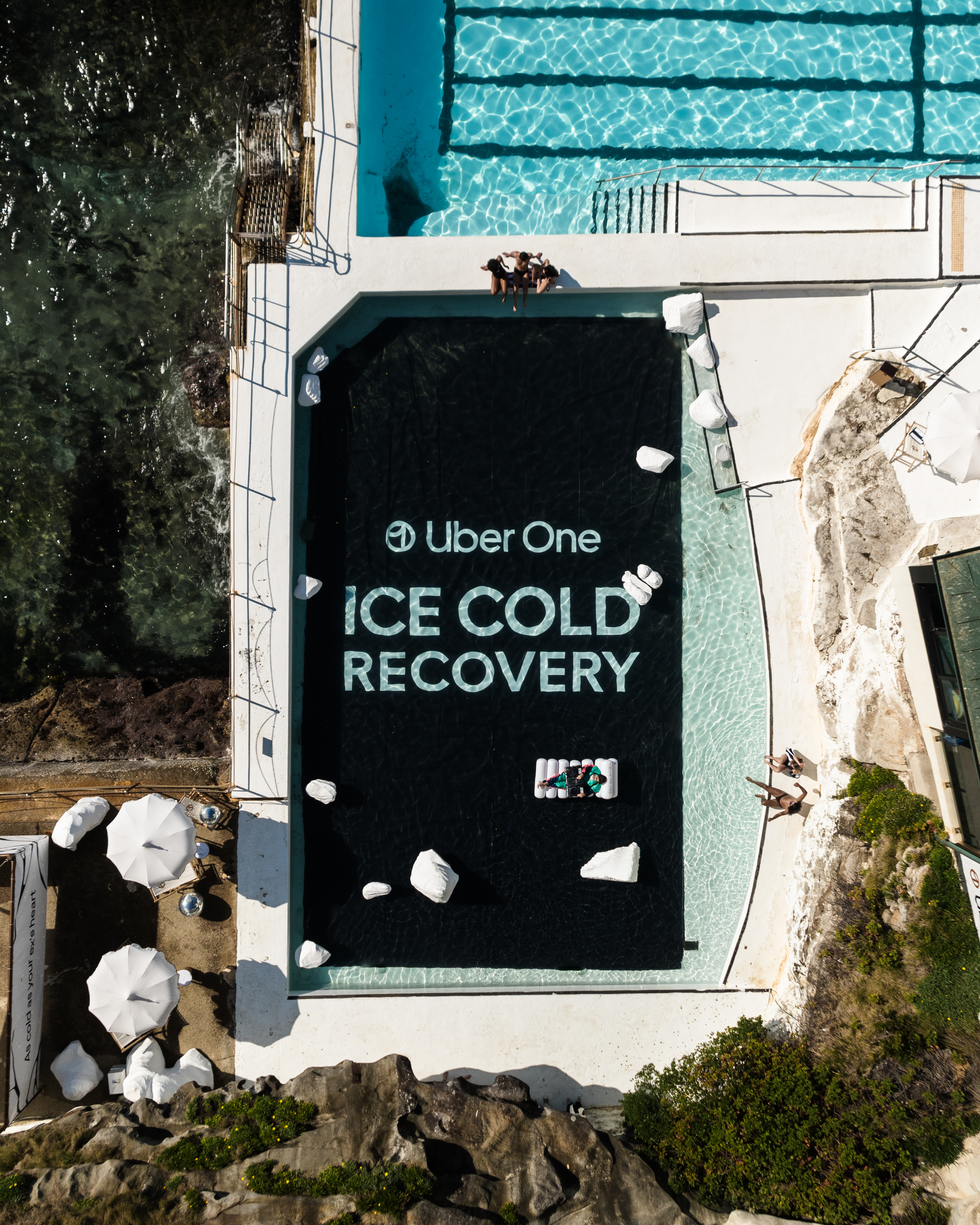 Aerial view of a pool with "Uber One Ice Cold Recovery" text, surrounded by white lounge areas and a coastline