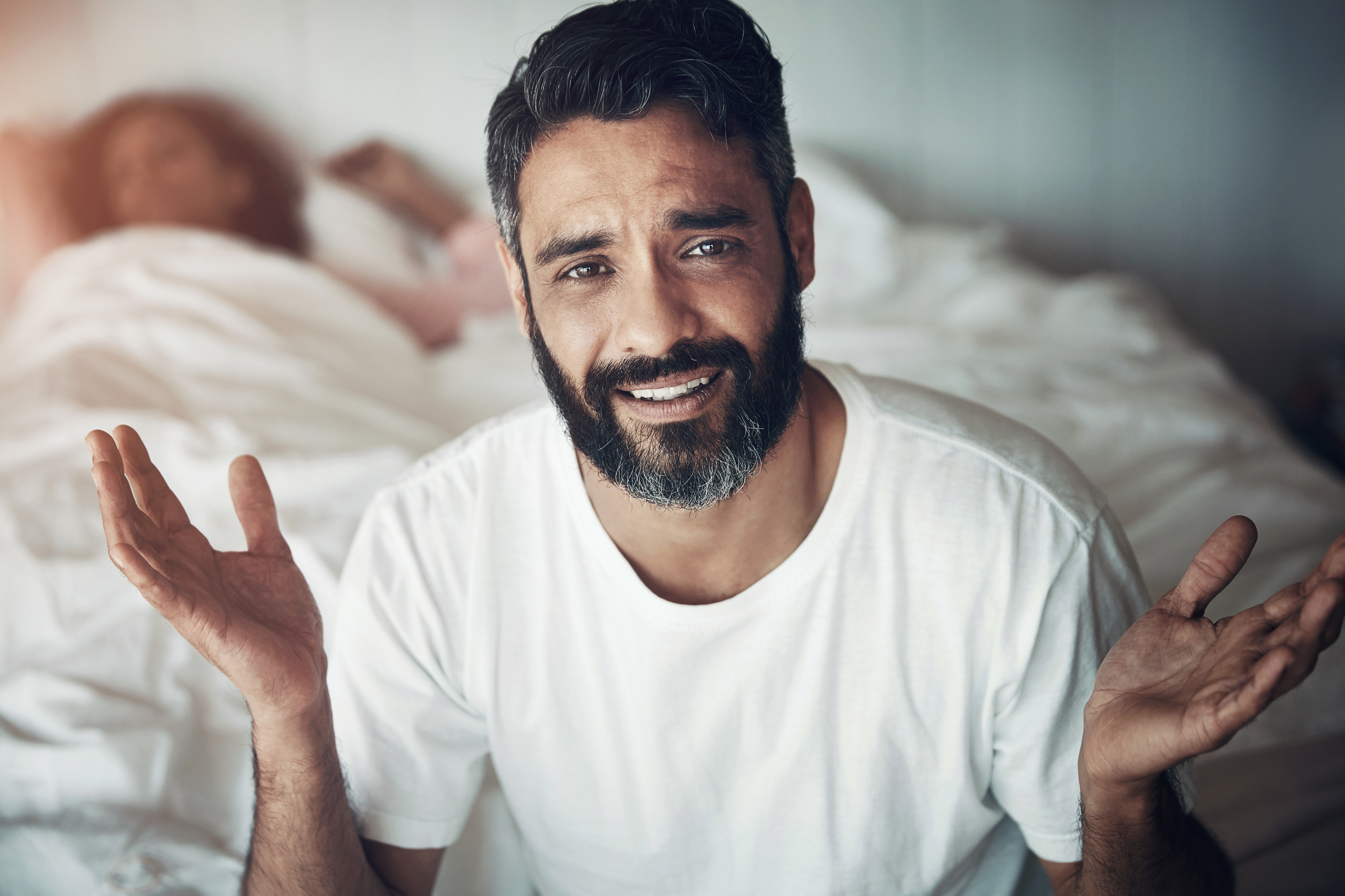 Man in a white t-shirt shrugs with a confused expression; a person sleeps in the bed behind him
