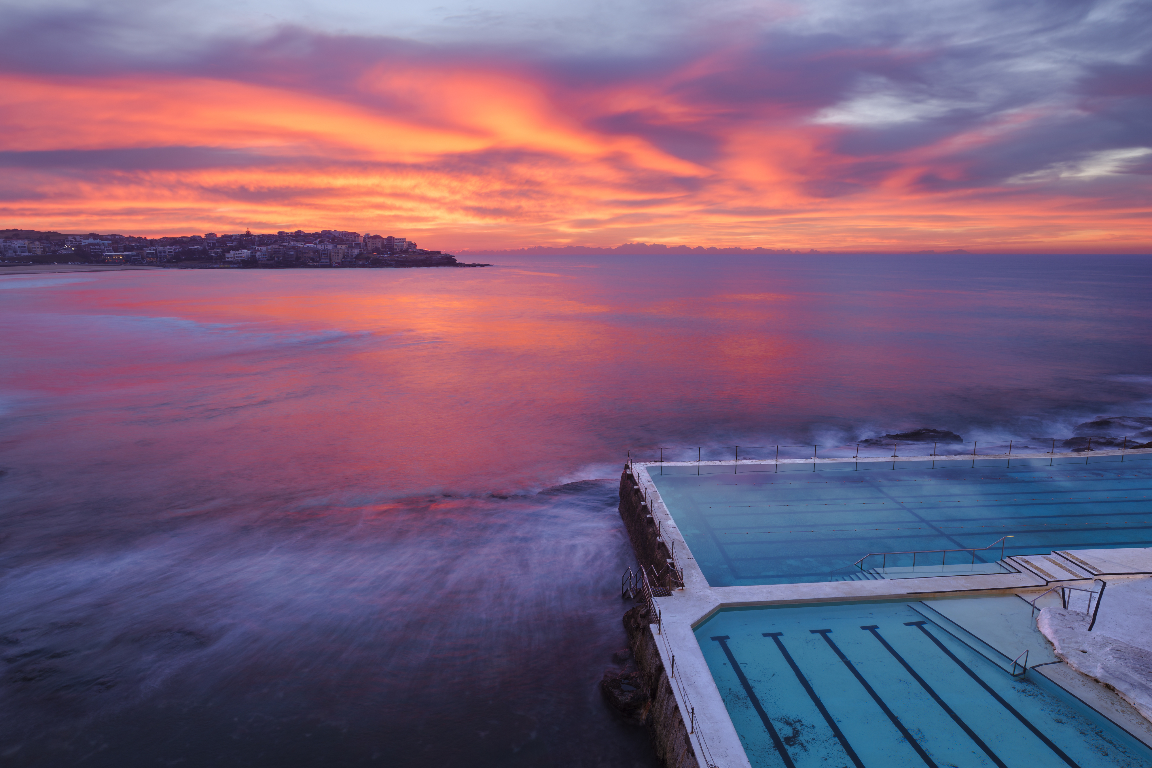 A beautiful ocean sunset scene with vibrant skies, overlooking a coastal infinity pool blending into the sea. The water is calm, and the horizon is distant