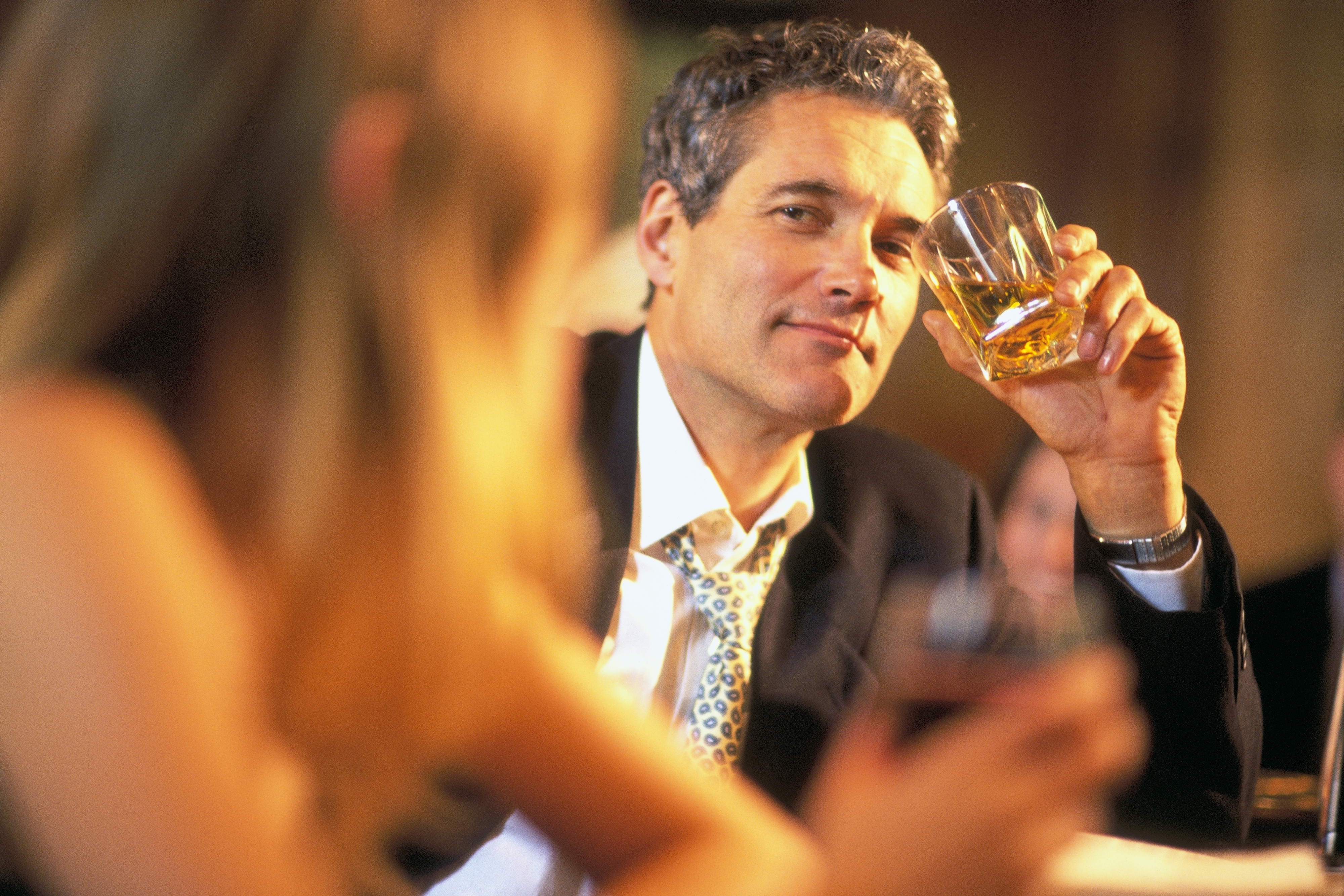 A man with a loosened tie holds a glass of whiskey while seated at a bar, smiling at a woman who is facing away