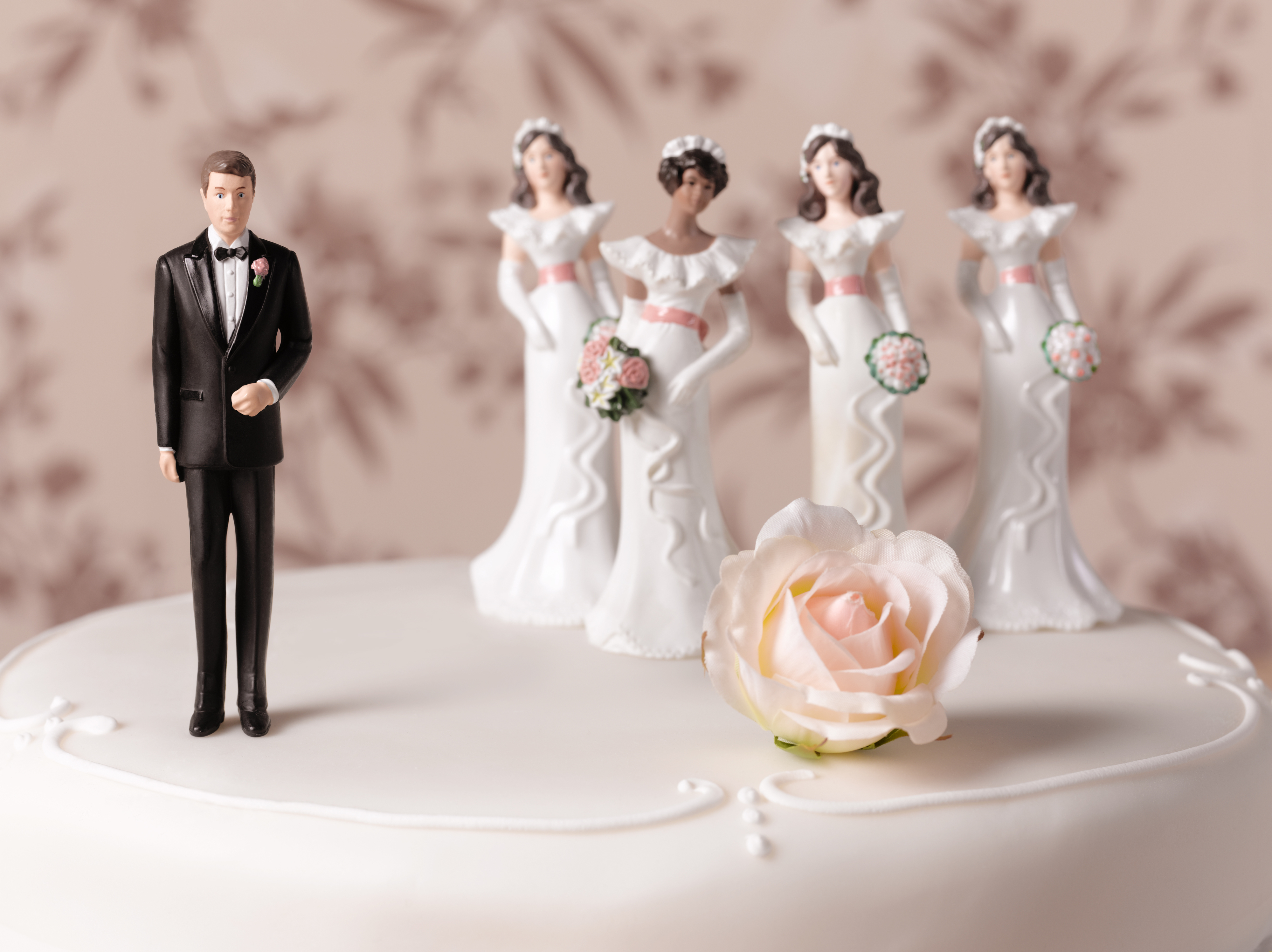 A wedding cake topper features a groom figurine next to four bride figurines holding bouquets, with a rose decoration on the cake