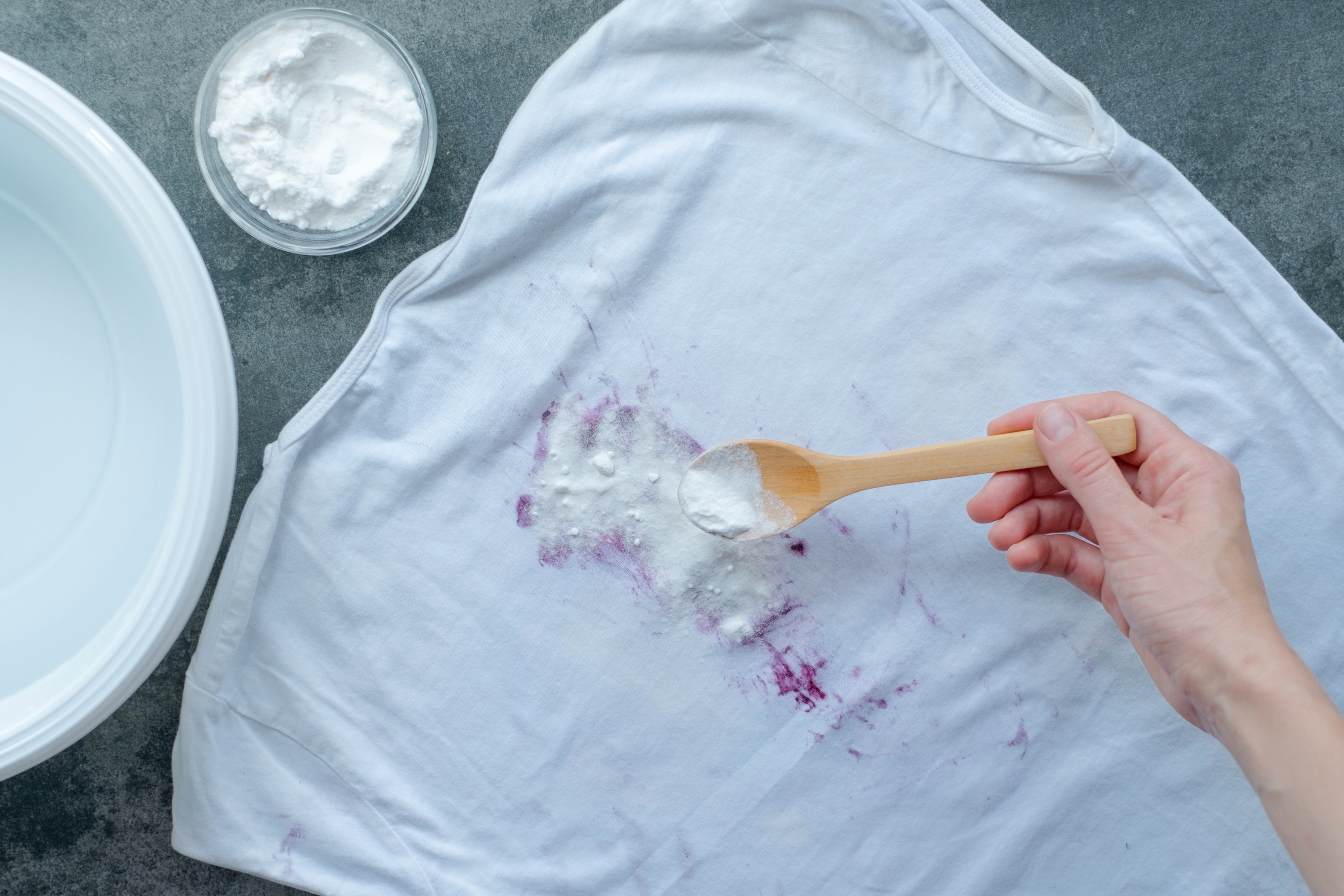 A hand uses a wooden spoon to apply powder to a stained white shirt, positioned near a washing basin and a small bowl of more powder