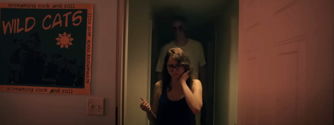 A scene from a movie shows a concerned woman walking through a dimly lit hallway with a shadowy figure in the background. A poster for &quot;Wild Cats&quot; is visible on the wall