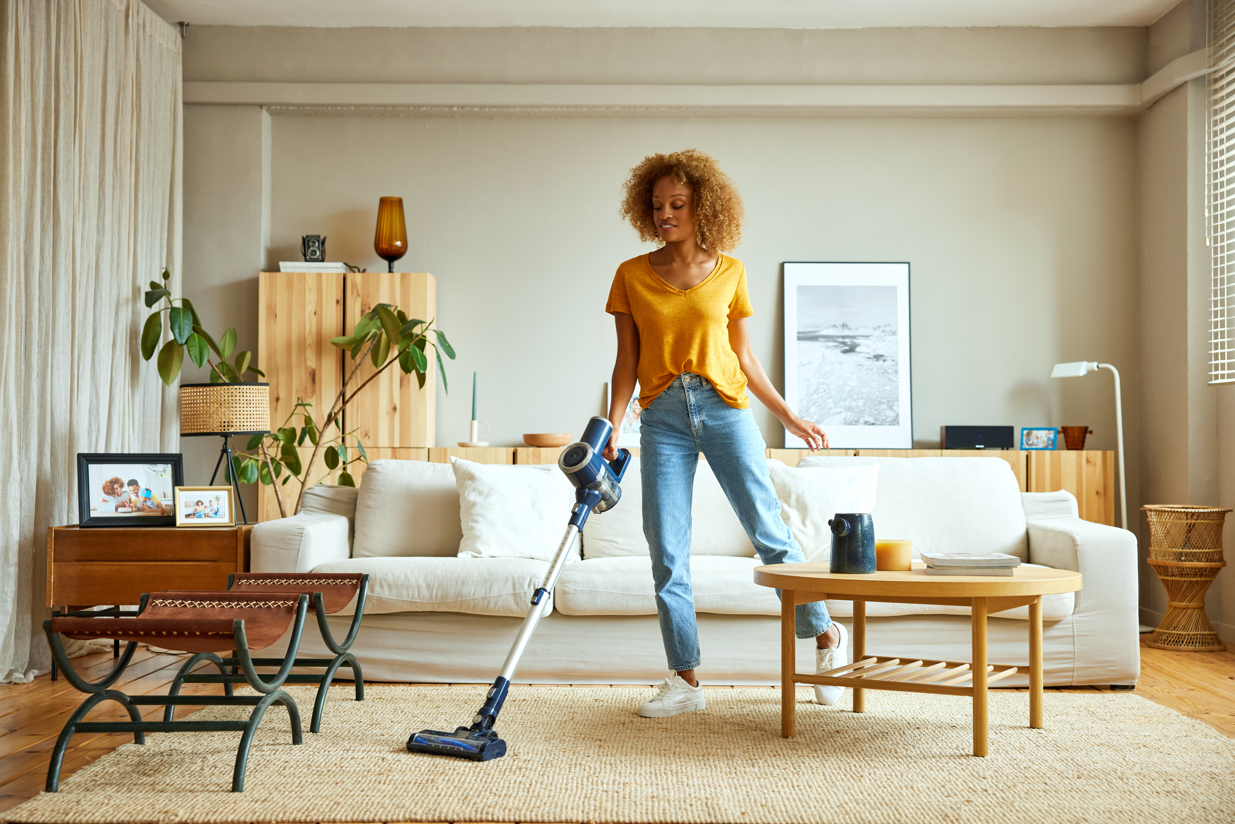 A woman vacuums her living room, which features a beige sofa, a wooden coffee table, and potted plants