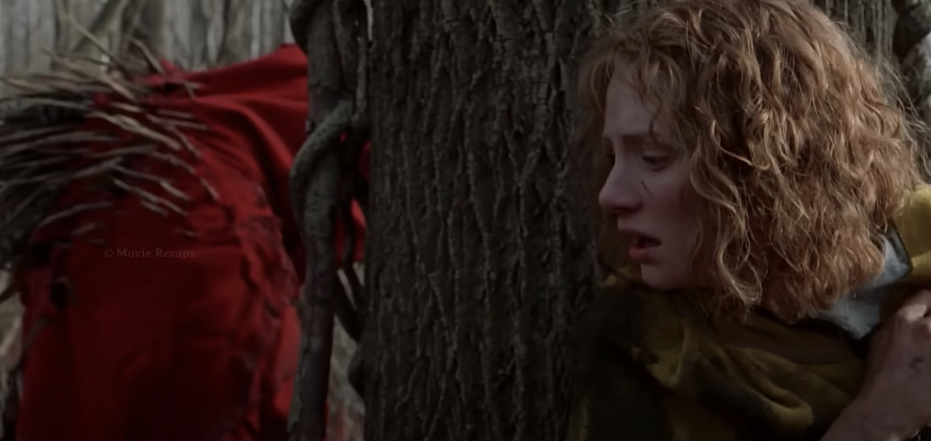 Bryce Dallas Howard, looking fearful, hides behind a tree in a forest while a menacing creature in red approaches in a scene from the film &quot;The Village.&quot;