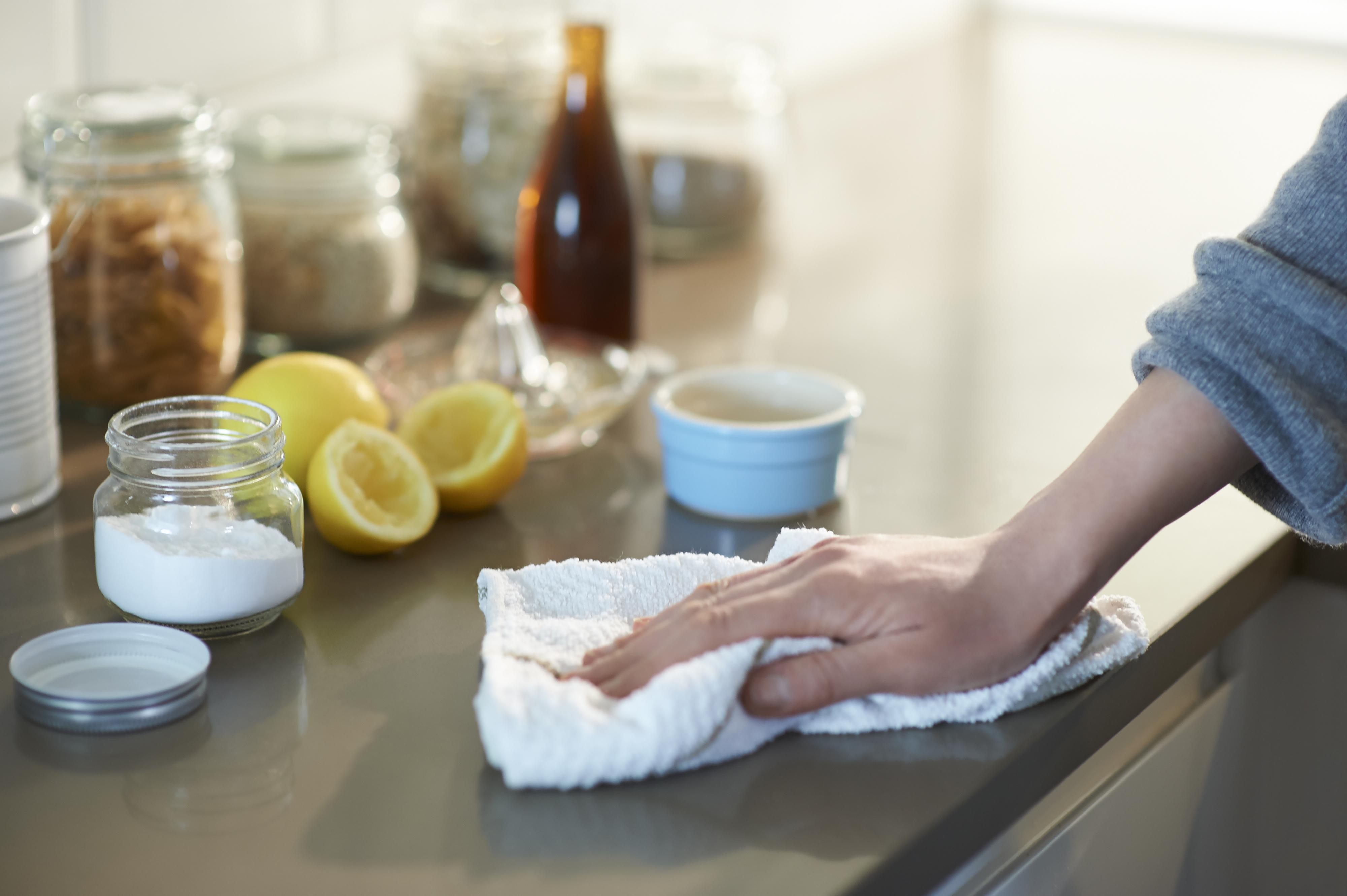 A hand wipes a kitchen counter with a cloth. Nearby are lemons, a small bowl, a jar of baking soda, and various containers