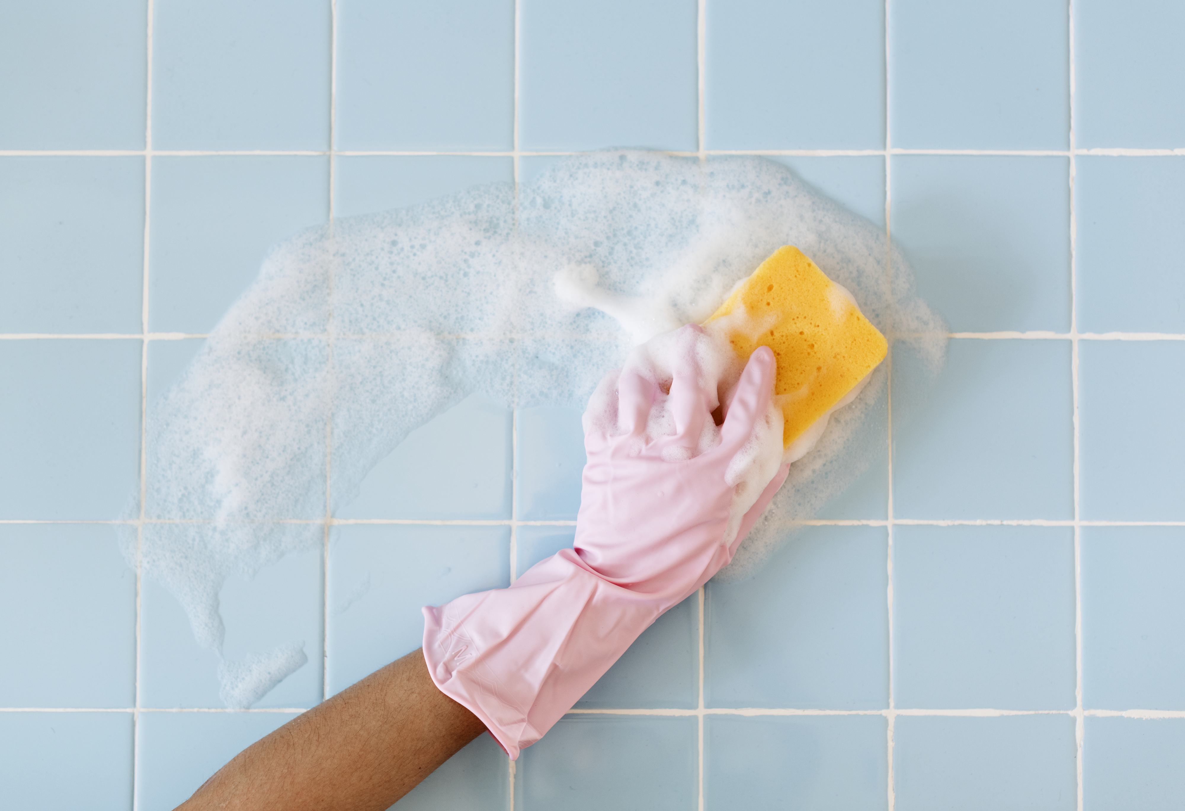 A gloved hand is scrubbing soapy tiles with a sponge