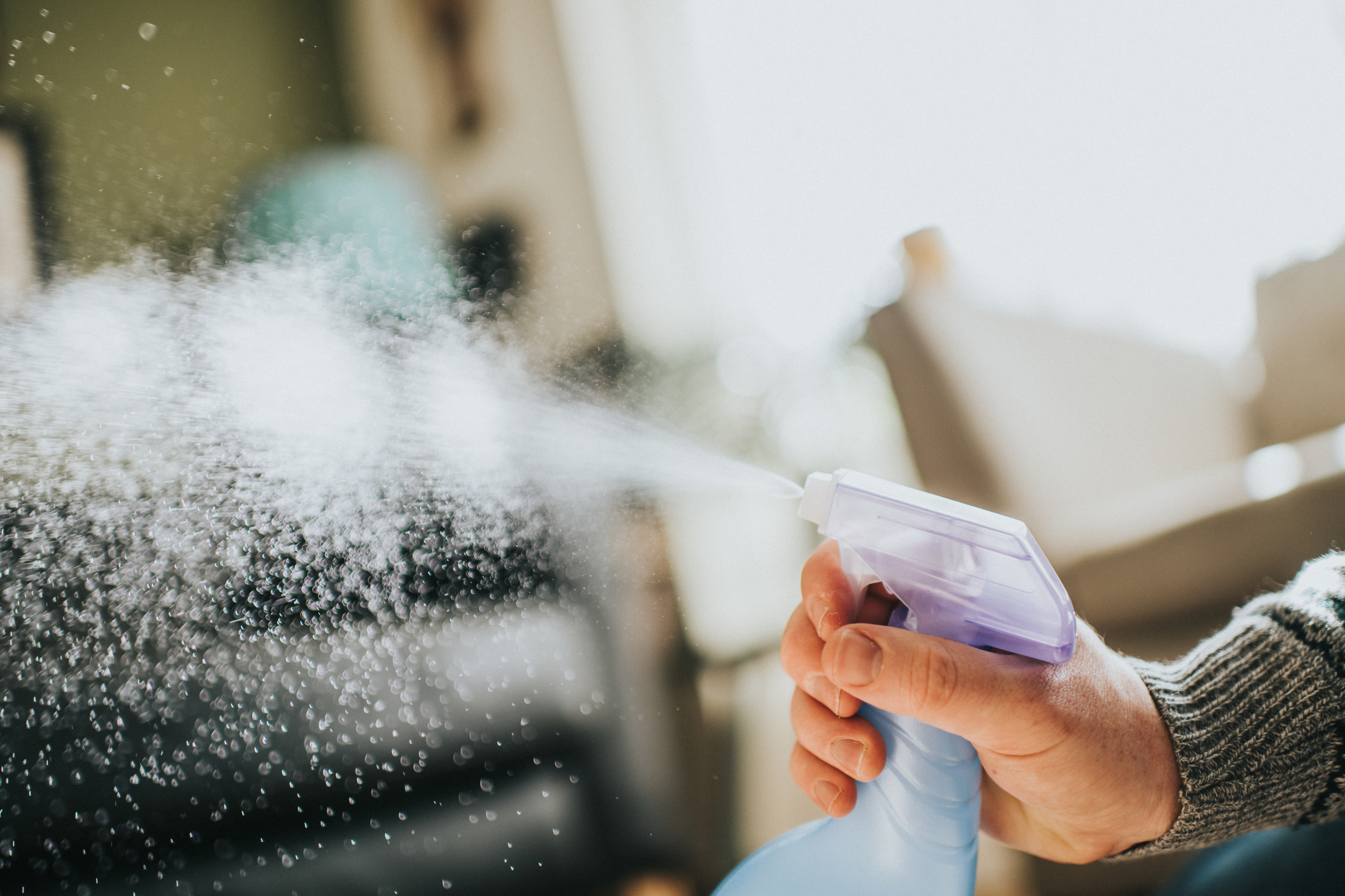 A hand holds a spray bottle, releasing a mist of liquid into the air inside a home
