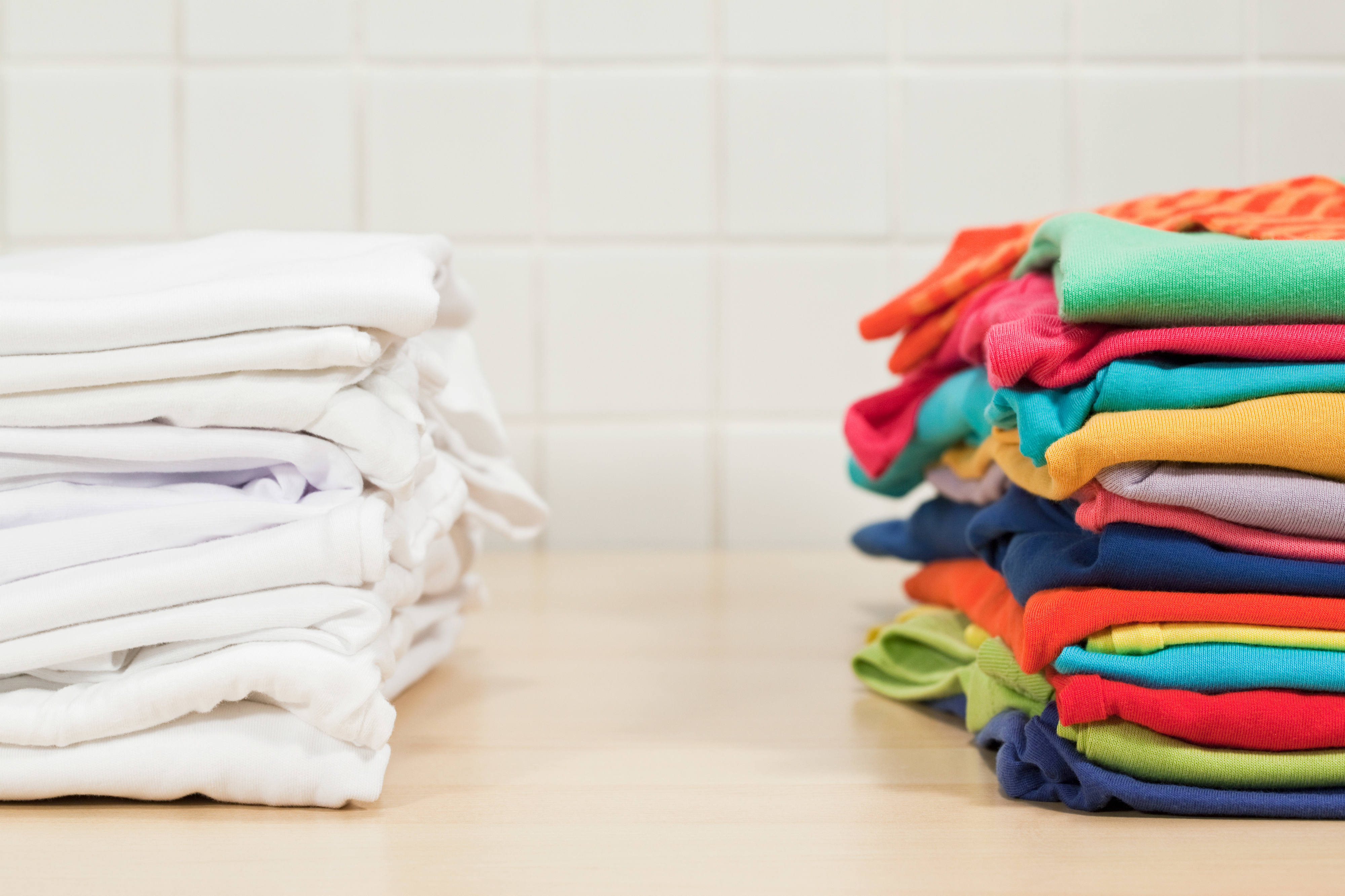 A stack of folded white clothes is on the left, while a stack of folded colorful clothes is on the right, both on a wooden surface