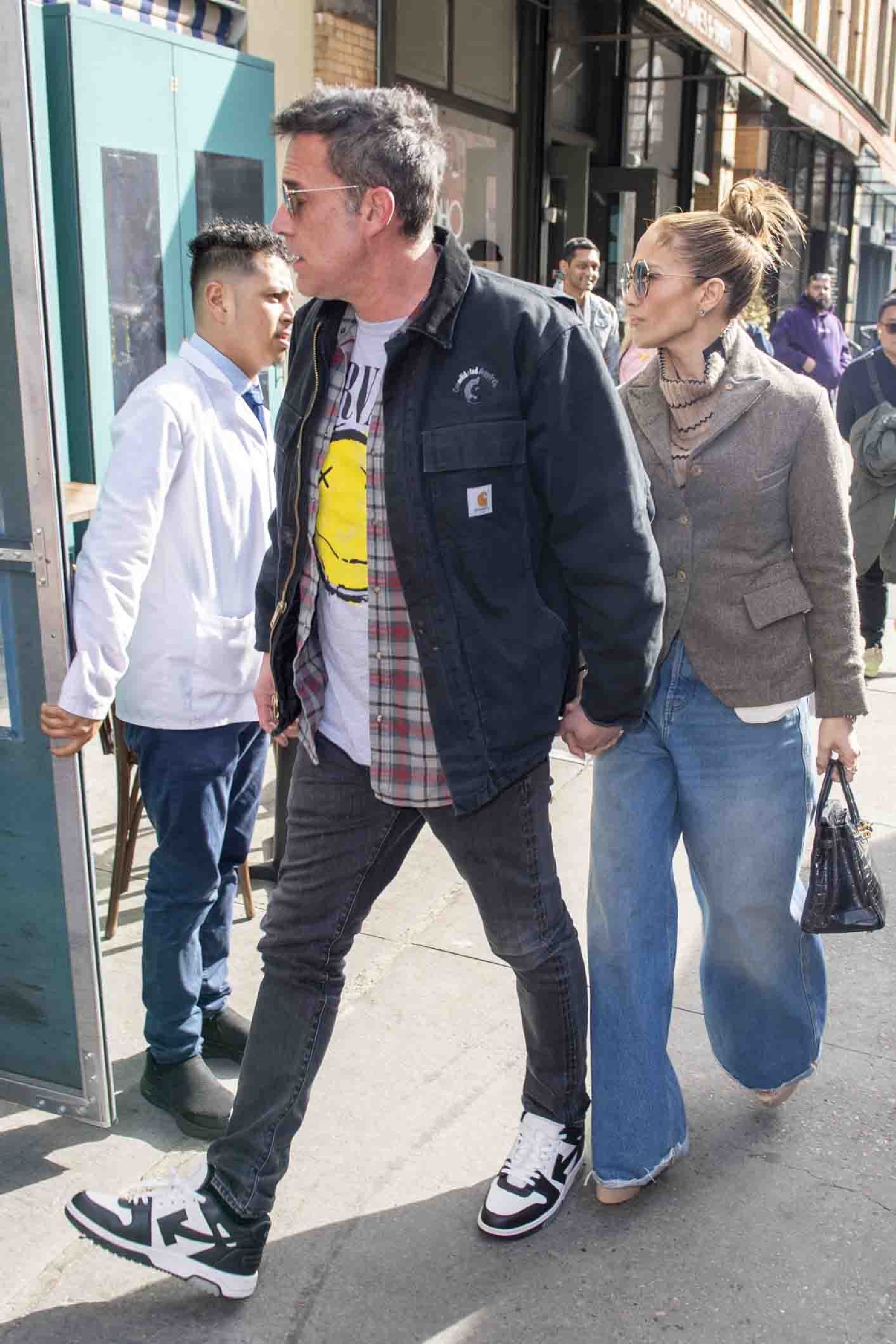 Ben Affleck and Jennifer Lopez hold hands walking outside. Ben wears a graphic tee, plaid shirt, dark jeans, and a jacket; Jennifer wears a gray blazer and wide-leg jeans