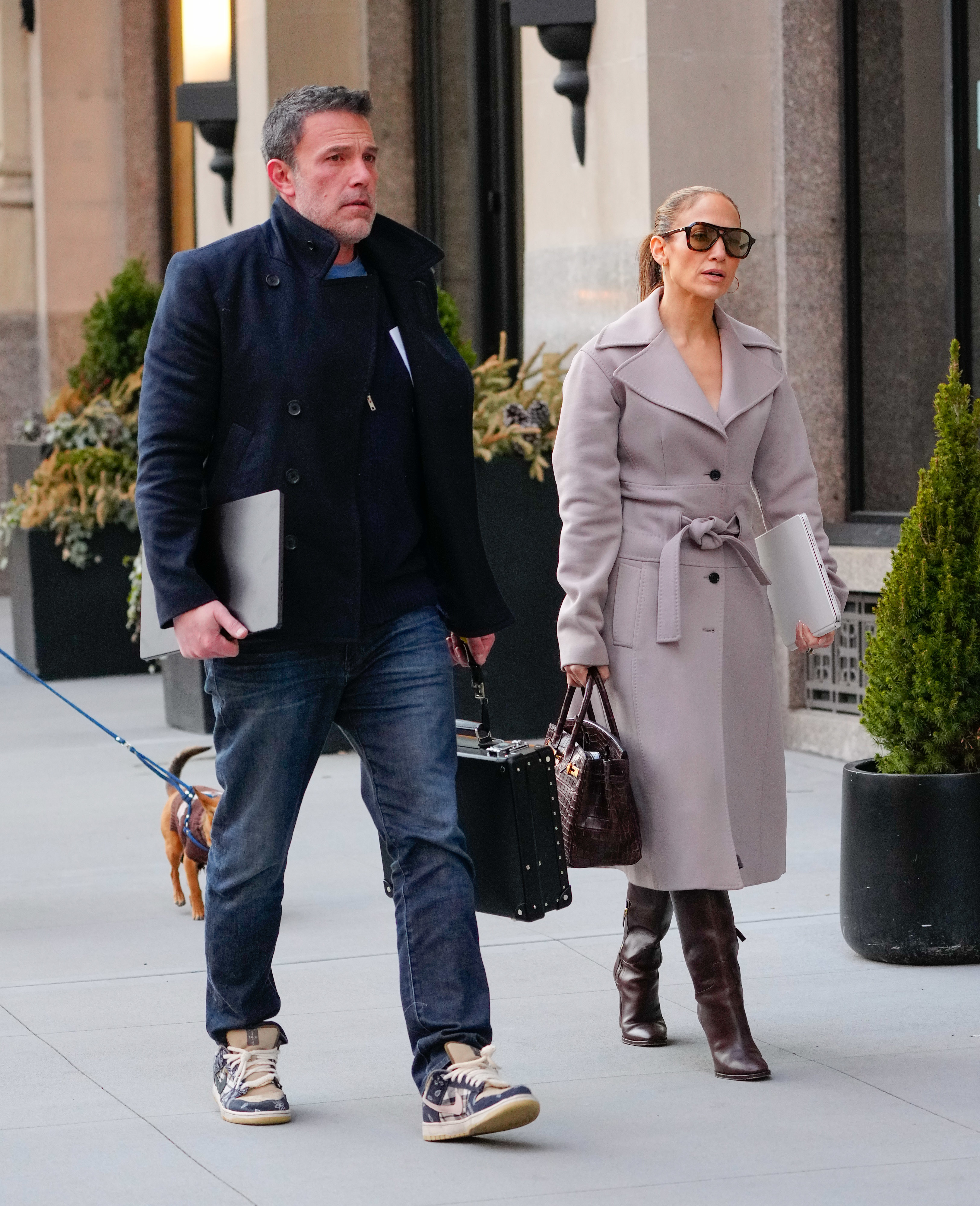 Ben Affleck and Jennifer Lopez walk on a sidewalk; Ben Affleck wears a coat and jeans, Jennifer Lopez wears a trench coat and knee-high boots, both holding laptops