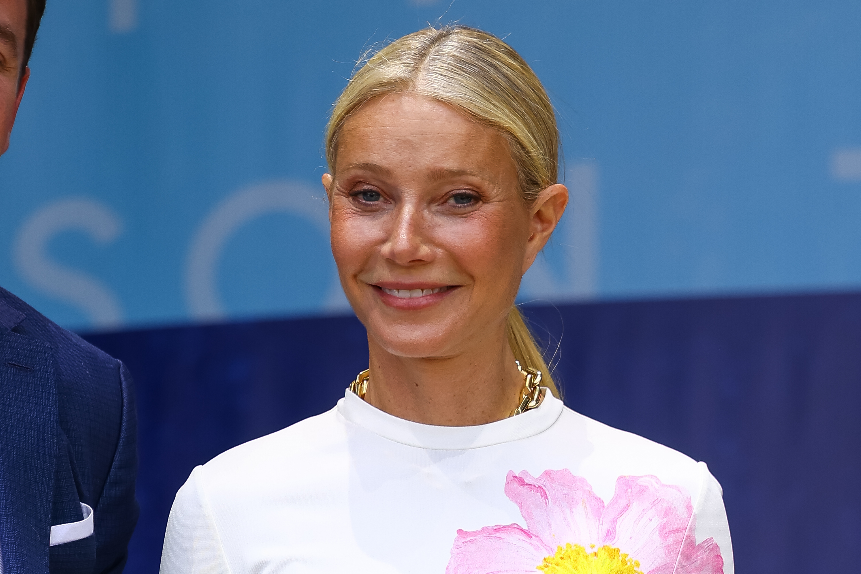 Gwyneth Paltrow is smiling while wearing a white outfit with a large pink floral design on the shoulder. She is in front of a blue background