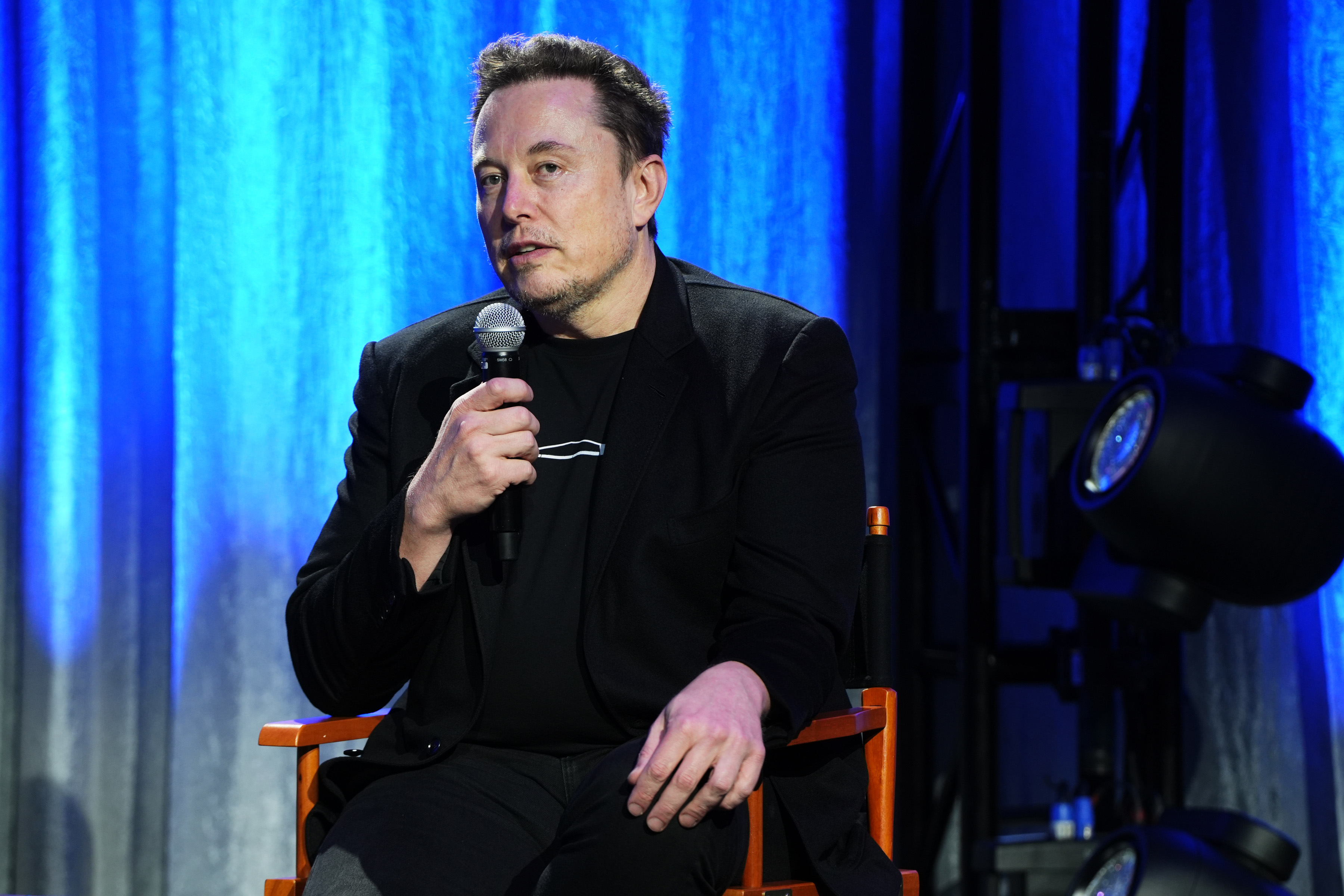 Elon Musk speaks into a microphone while seated on stage, wearing a black blazer and black shirt. Blue curtains hang in the background