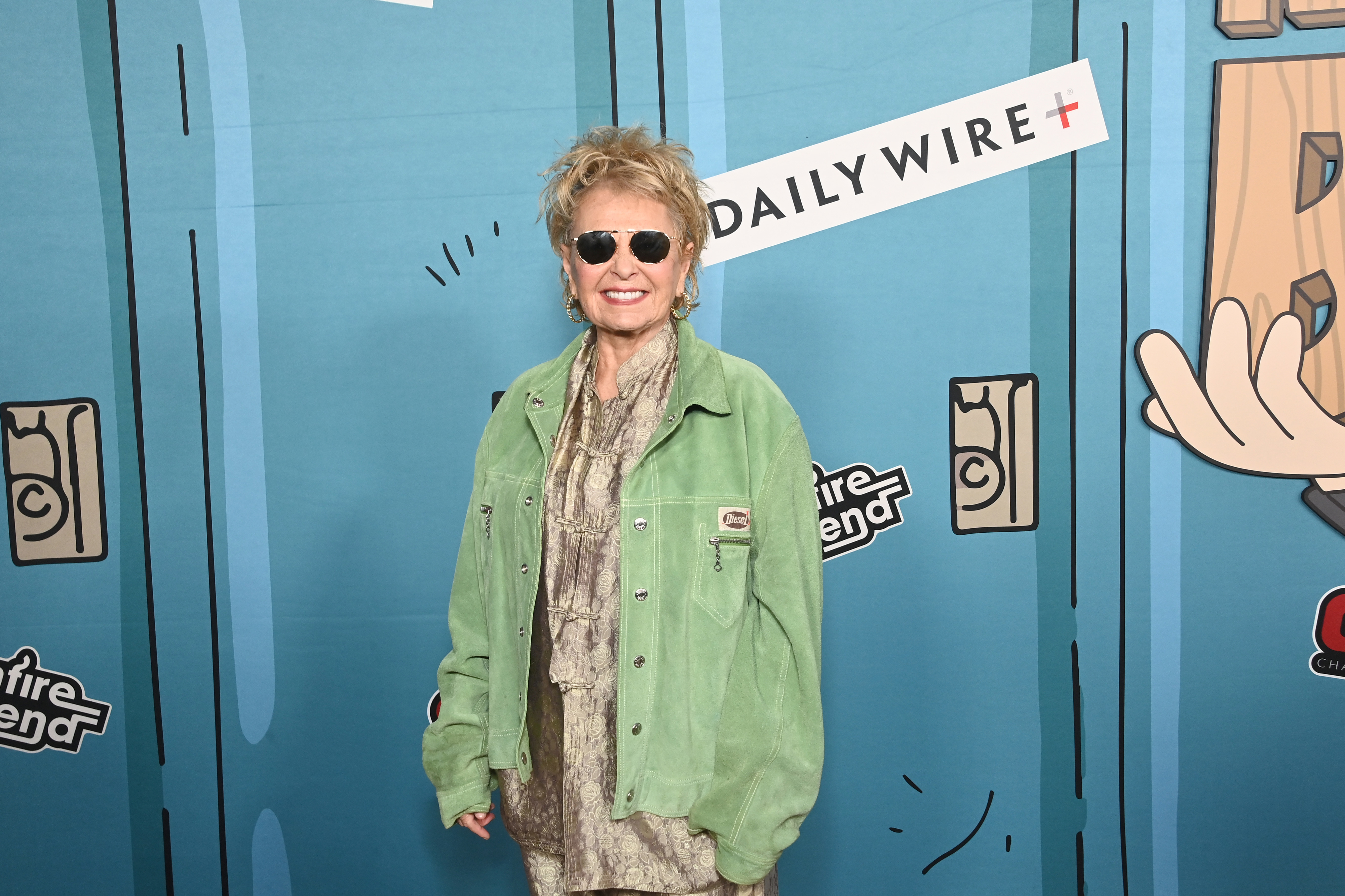 Roseanne Barr wearing sunglasses and a green jacket attends a Daily Wire event. She is standing in front of a cartoonish backdrop