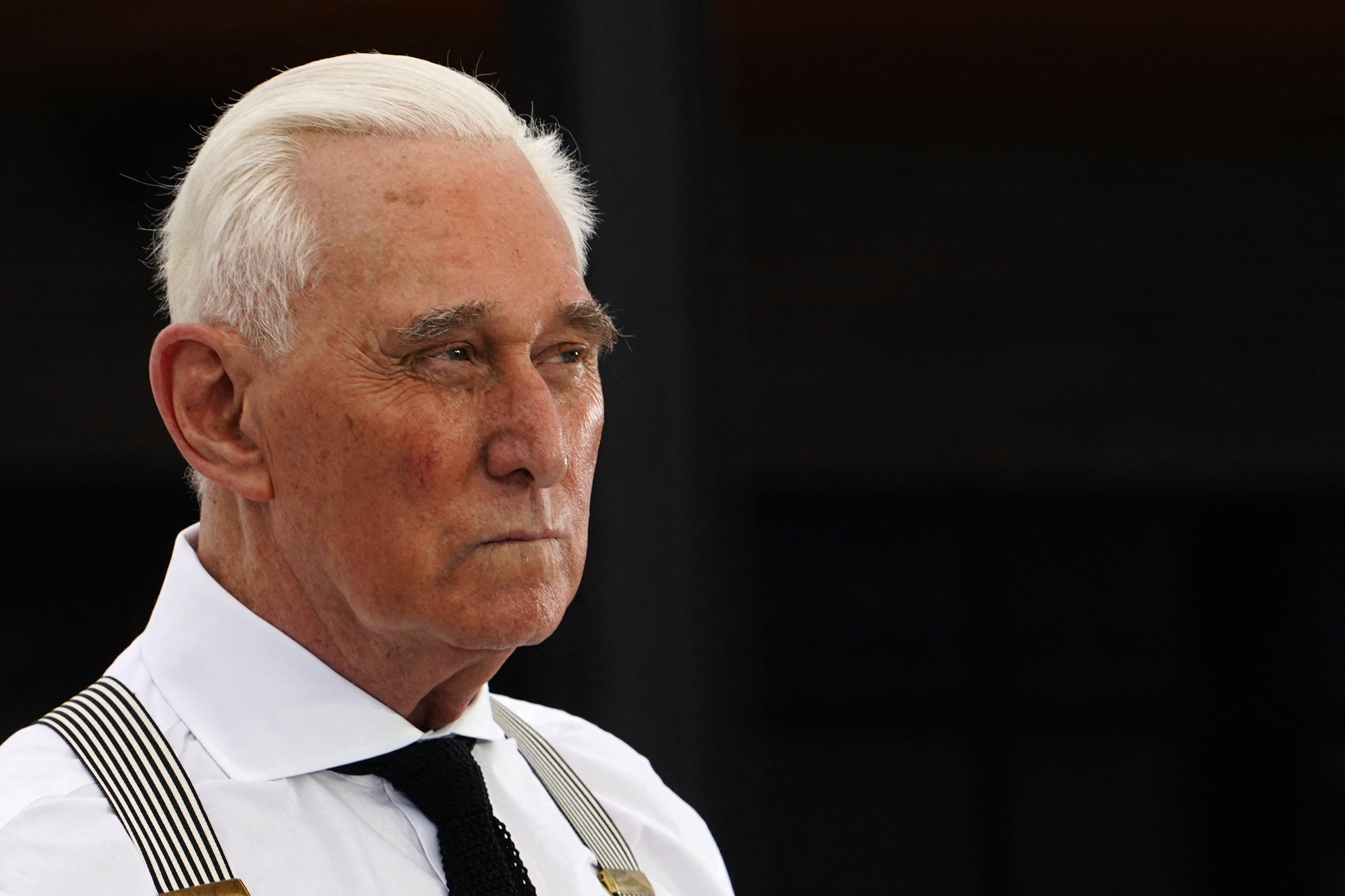 Roger Stone is wearing a white dress shirt with striped suspenders. He has a stern expression and is looking to the side