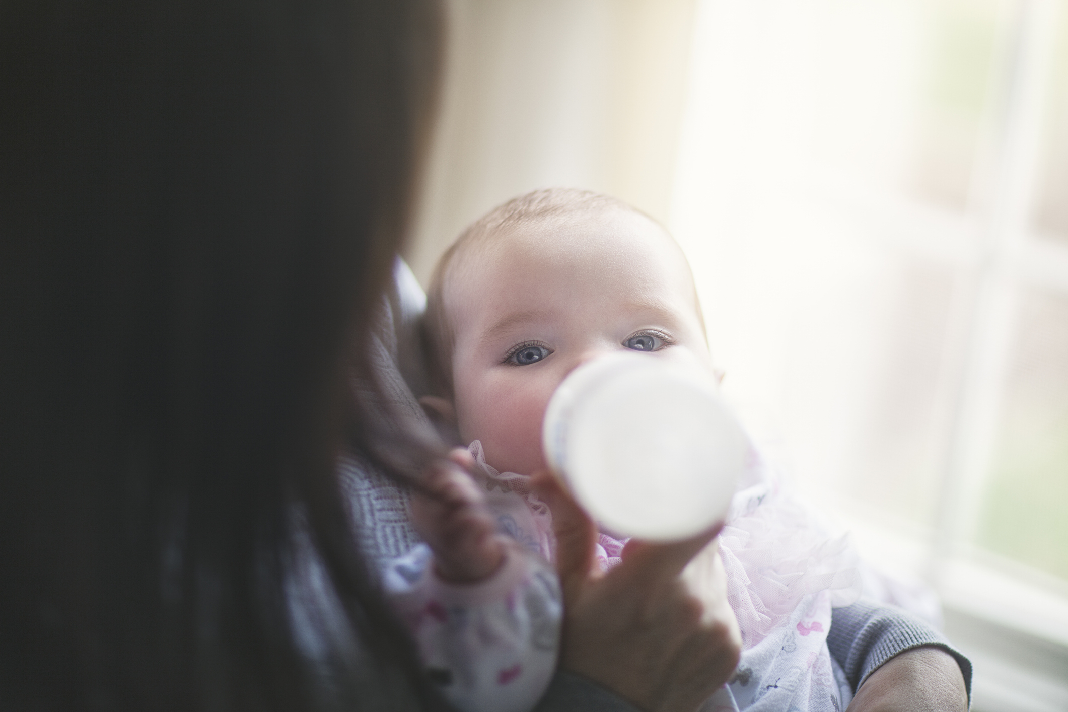 A baby is being fed with a bottle by an adult holding them. The baby looks towards the camera