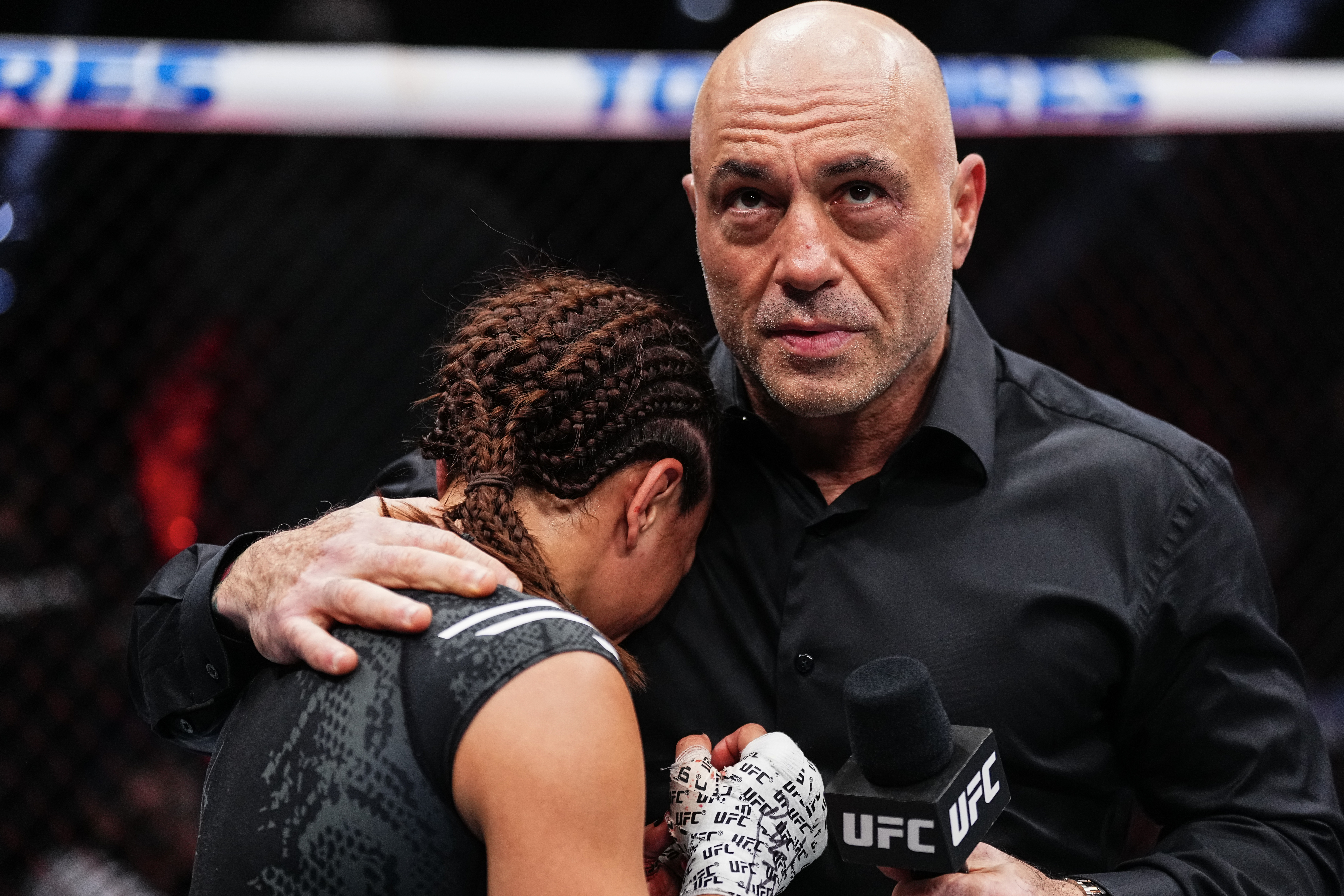 Joe Rogan consoles a female UFC fighter with braids in the octagon after a match, holding a UFC microphone