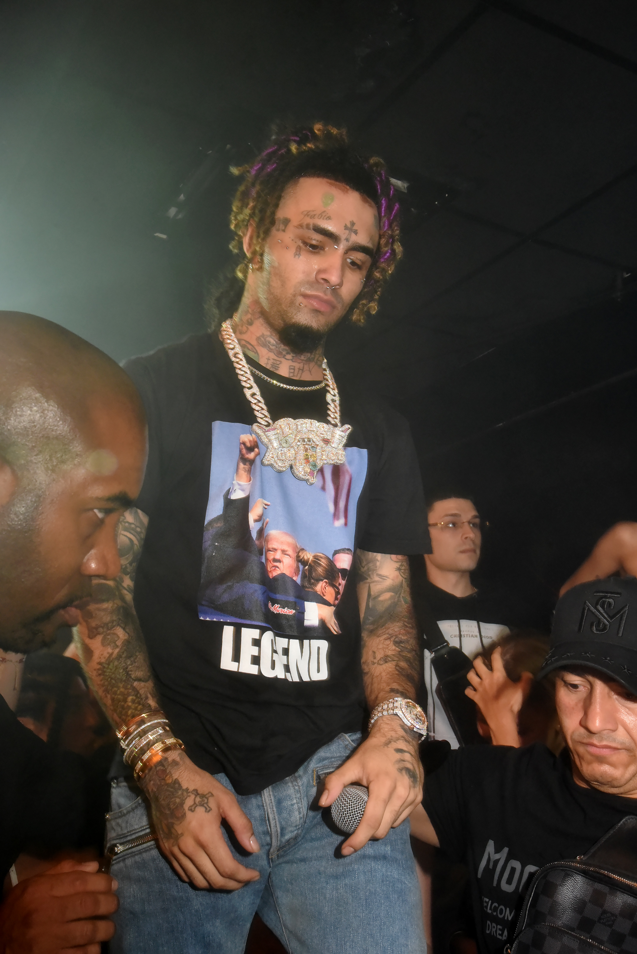 Lil Pump performing on stage, wearing a &quot;LEGEND&quot; t-shirt, jeans, and multiple gold chains. His signature face tattoos and bracelets are visible as he holds a microphone