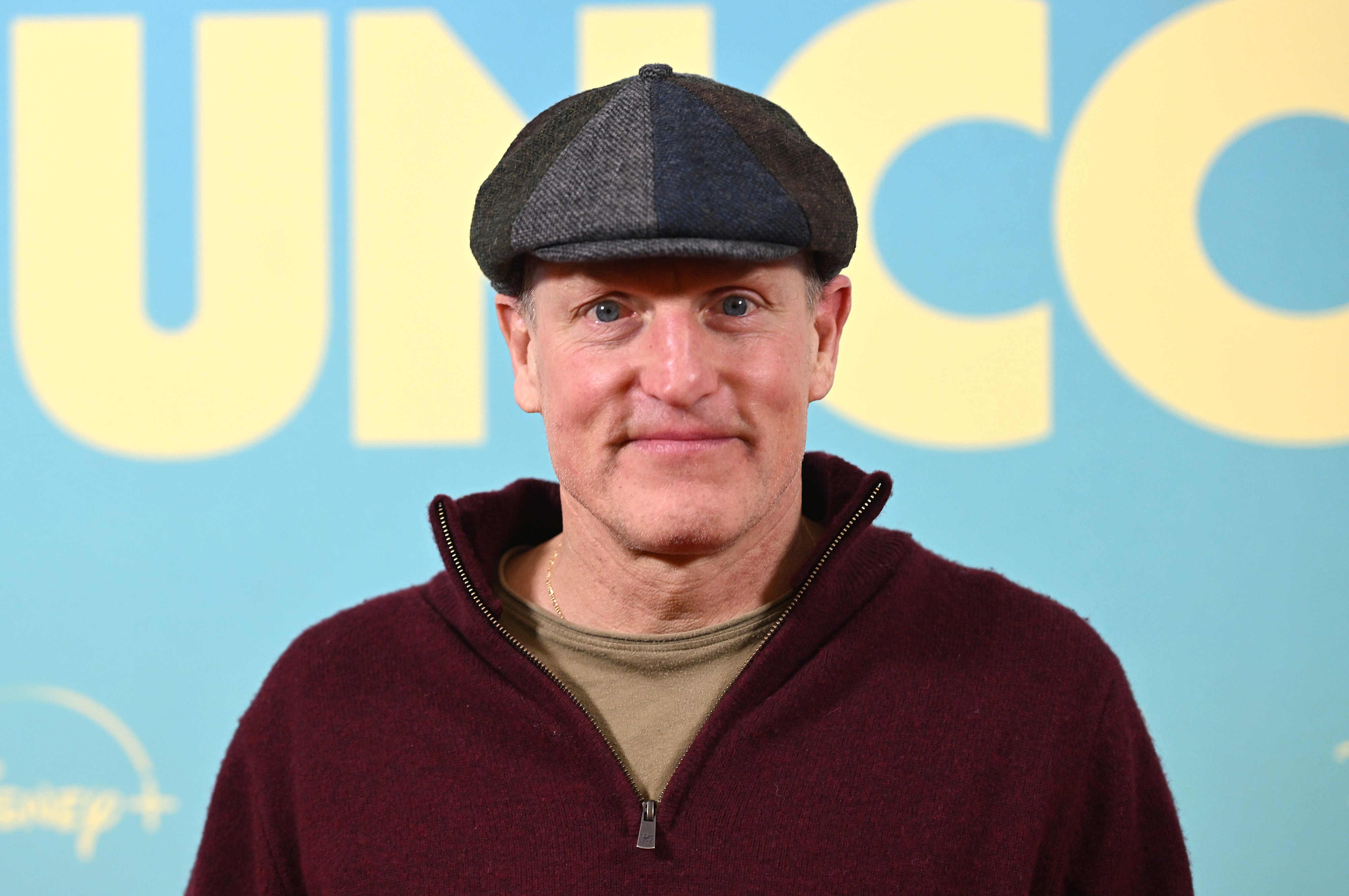 Woody Harrelson in a flat cap and zip-up jacket, standing in front of a backdrop with large text