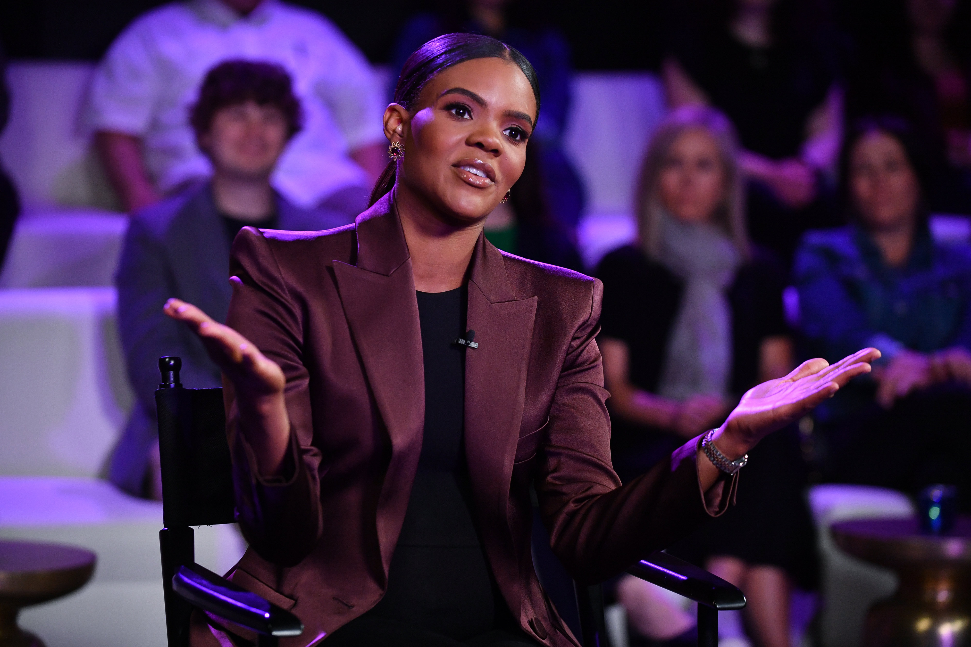 A woman sits in a chair, wearing a brown blazer with a black top, gesturing with her hands, smiling, and speaking to an audience in the background