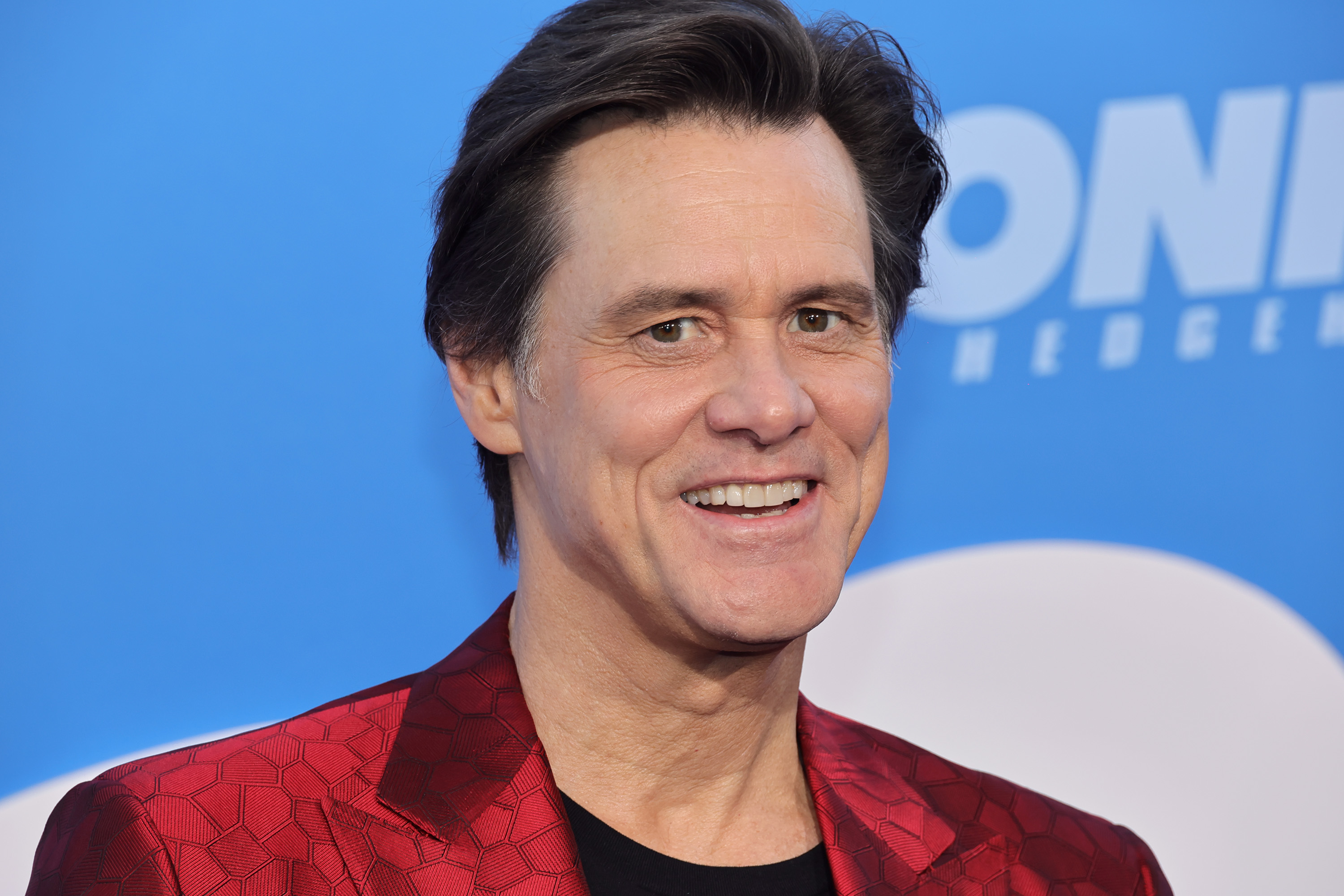 Jim Carrey at an event, smiling, wearing a red patterned jacket and black shirt, in front of a blue background with partial text visible