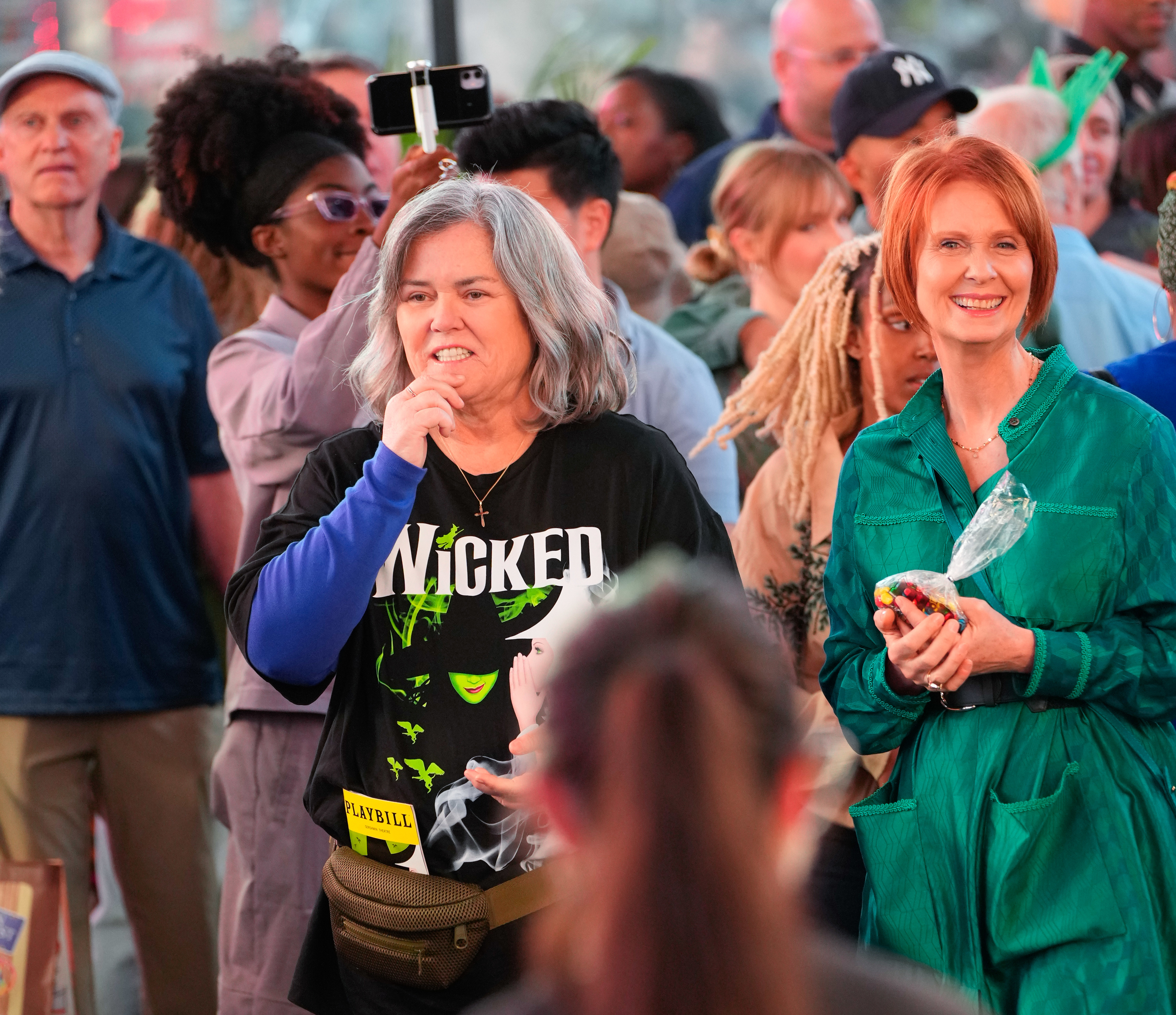 Rosie O&#x27;Donnell in a &quot;Wicked&quot; t-shirt and Cynthia Nixon in a green outfit are standing together in a crowded outdoor area