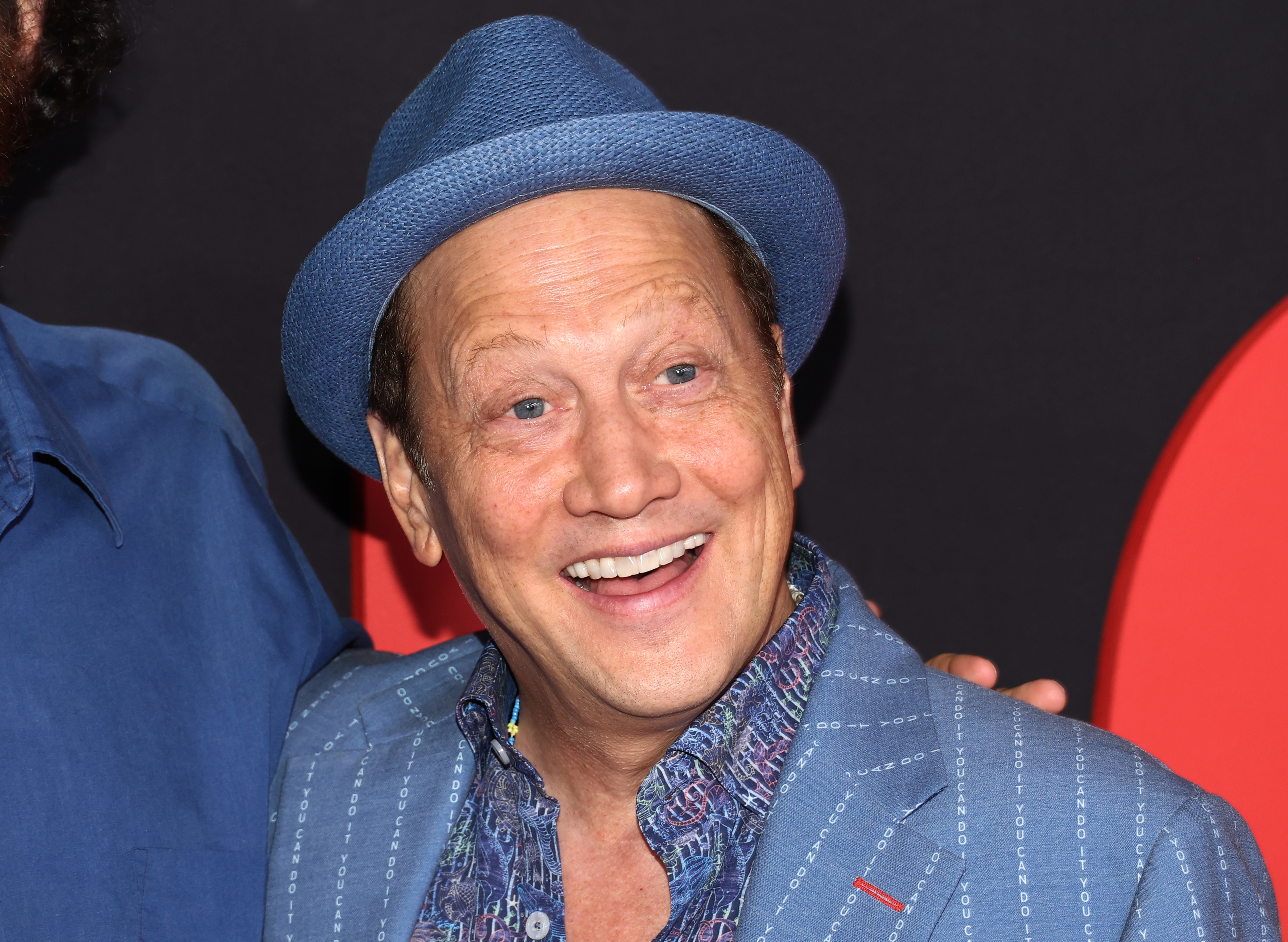 Rob Schneider smiles, wearing a patterned shirt and blue blazer with a matching blue fedora against a black and red background