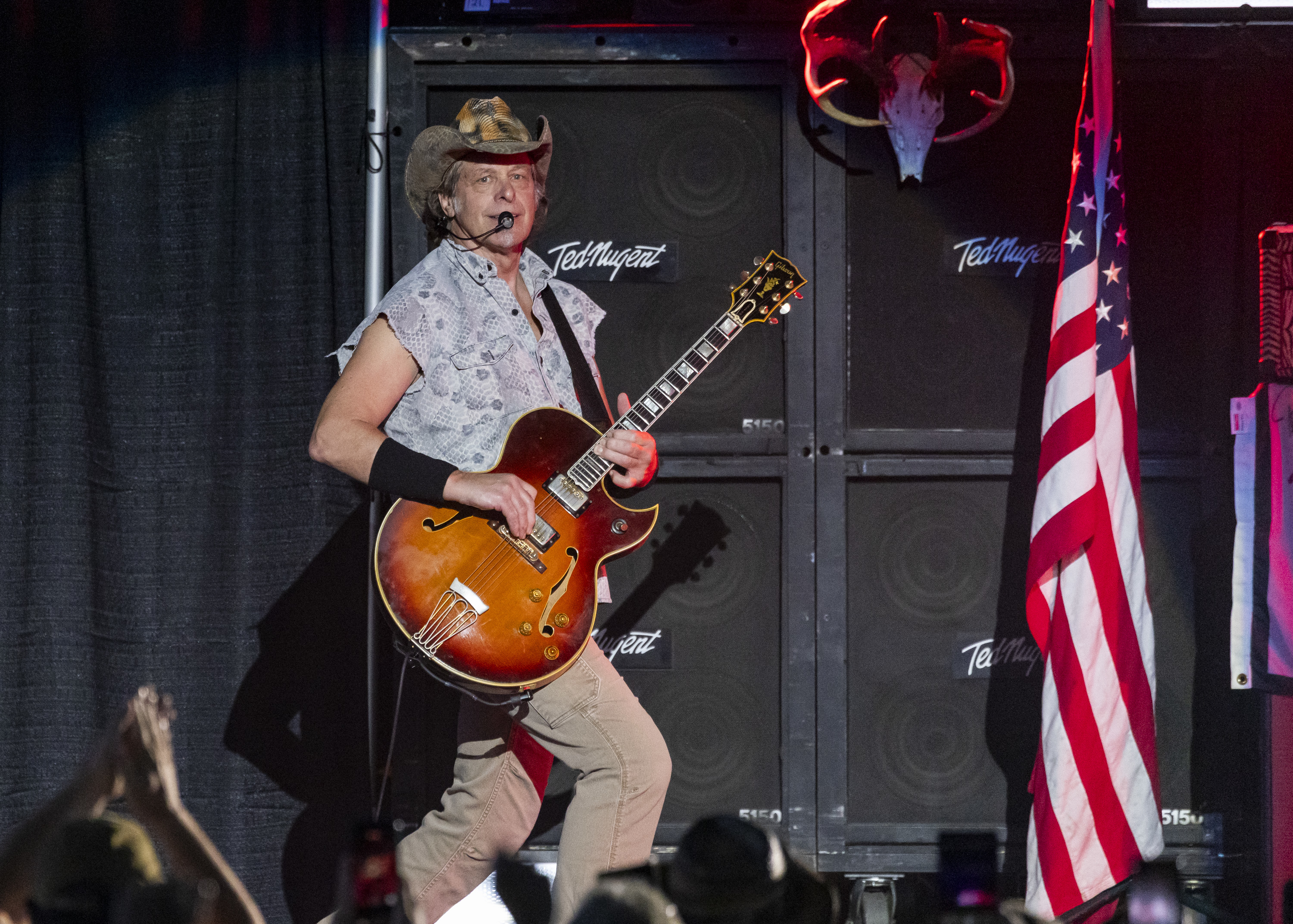 Ted Nugent performing on stage with an electric guitar, wearing a cowboy hat and a short-sleeved shirt. An American flag and speakers are visible in the background