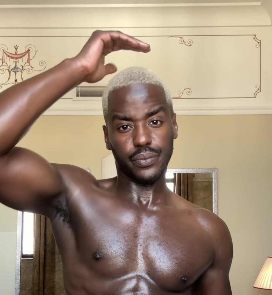 Ncuti Gatwa is shirtless, facing the camera with his hand on his head in an indoor setting