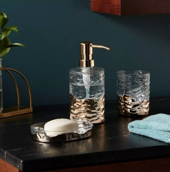 Elegant bathroom setup featuring a gold-trimmed glass soap dispenser, glass tumbler, soap dish with soap, teal towel, and a small plant on a wooden counter