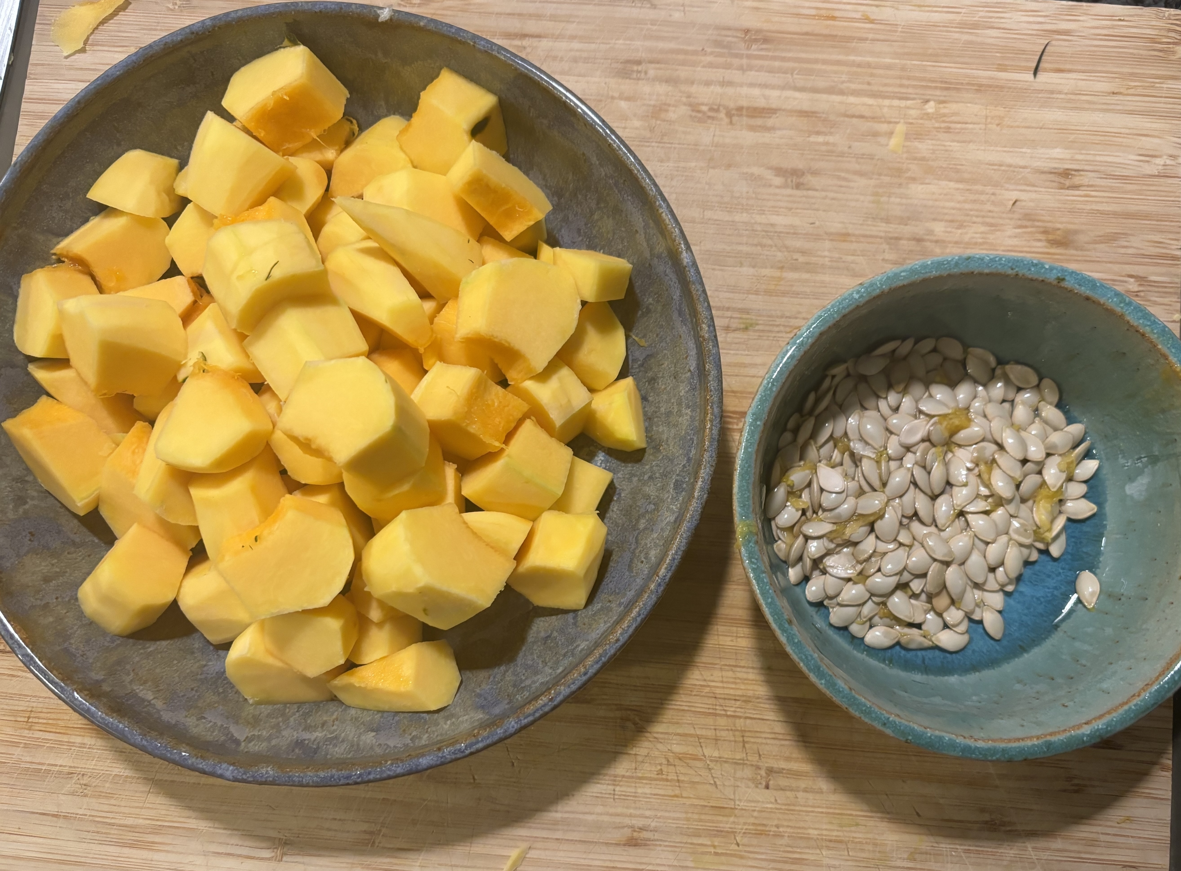 A bowl of diced butternut squash next to a bowl of butternut squash seeds on a wooden cutting board