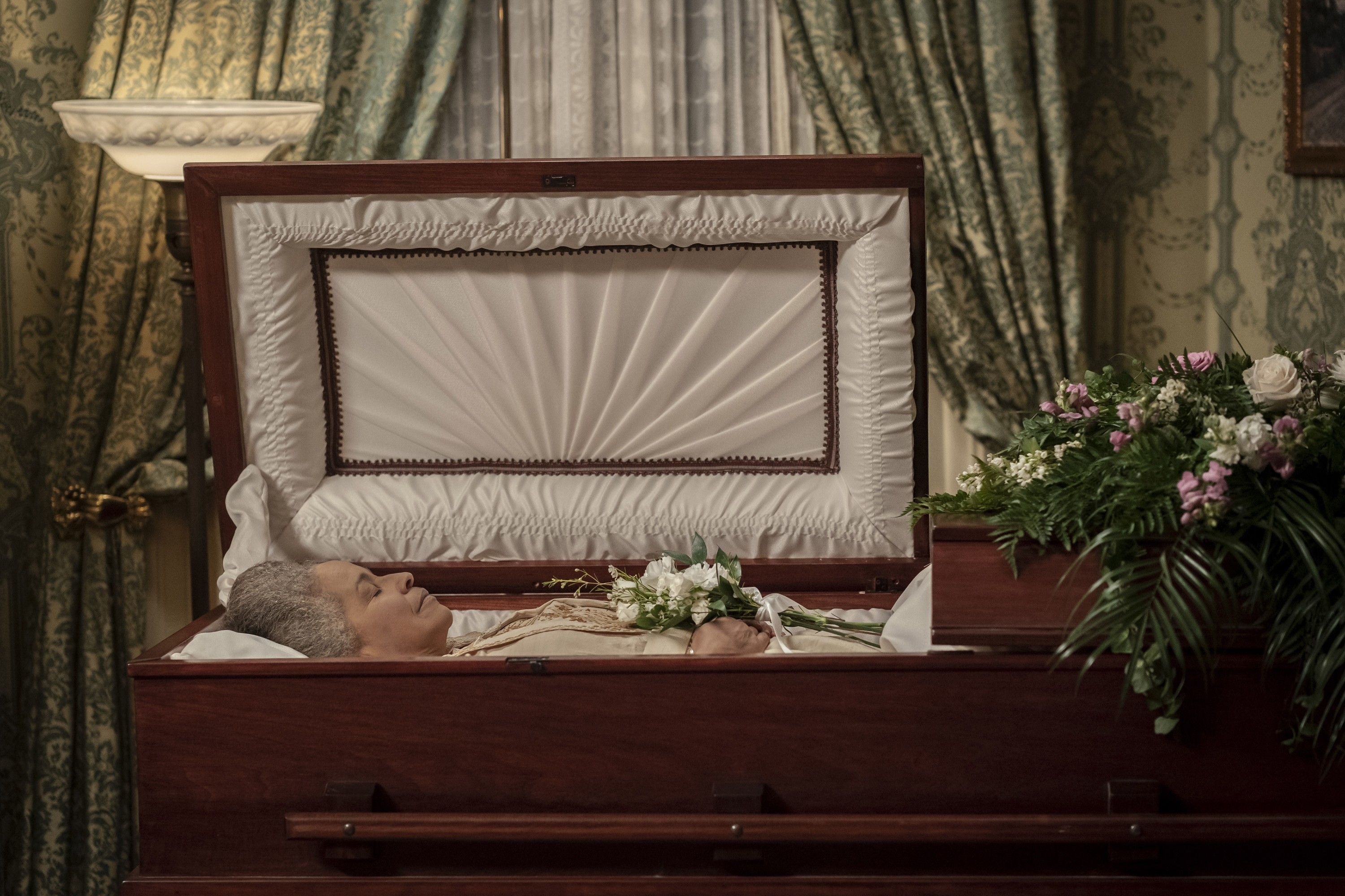 An elderly person is lying in a casket at a funeral service with floral arrangements on top of the casket