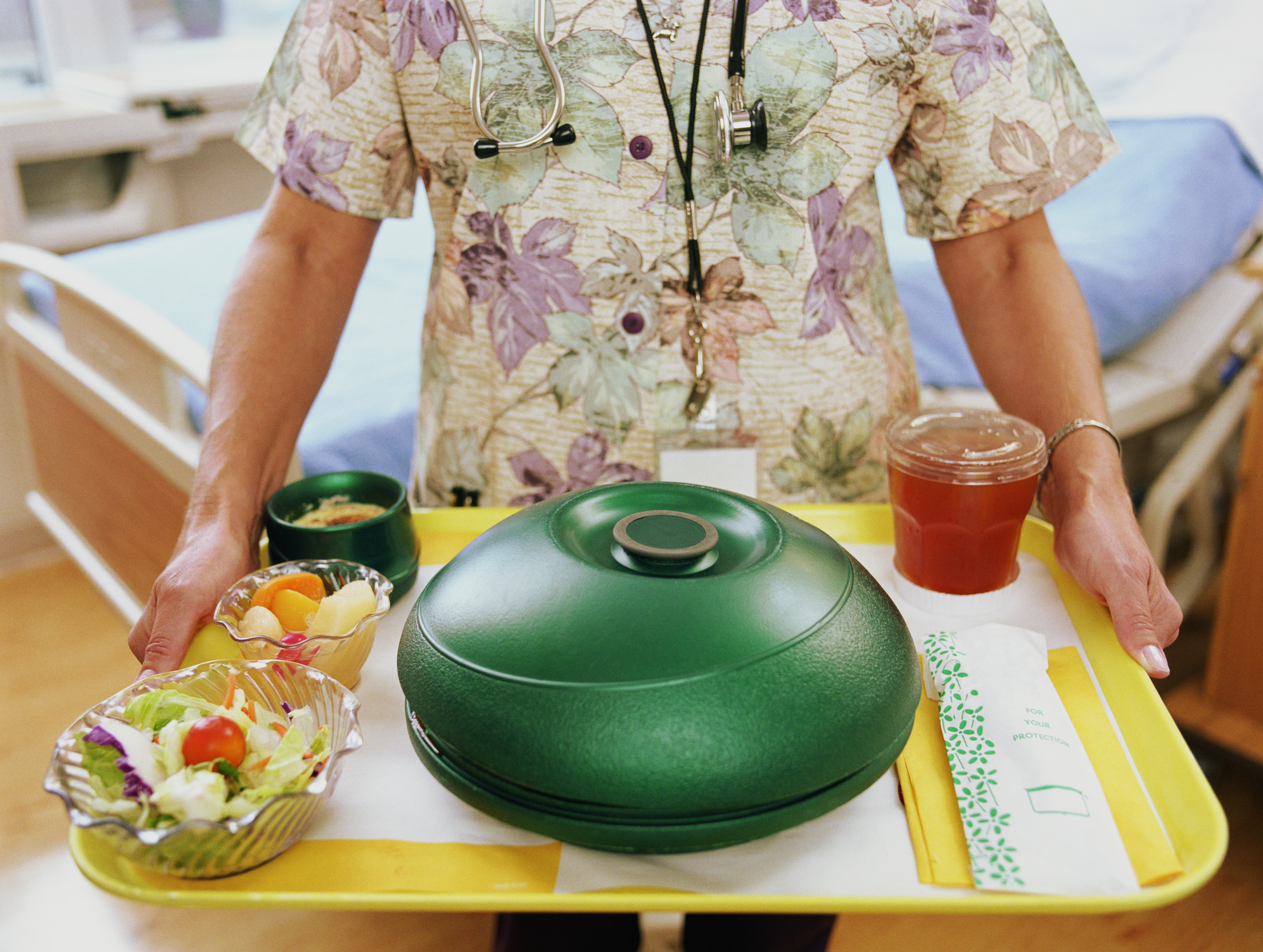 Person in patterned medical scrubs carries a hospital food tray with salad, fruit, soup, covered dish, drink, and utensils in a patient room