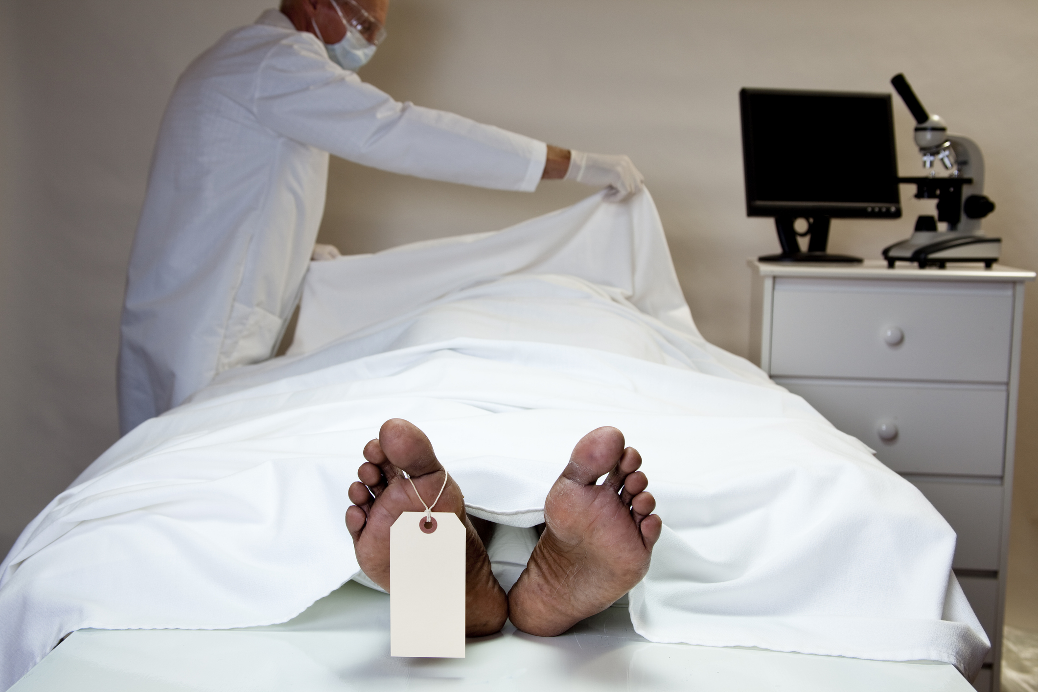 A medical examiner uncovers a body on an autopsy table, with a toe tag visible