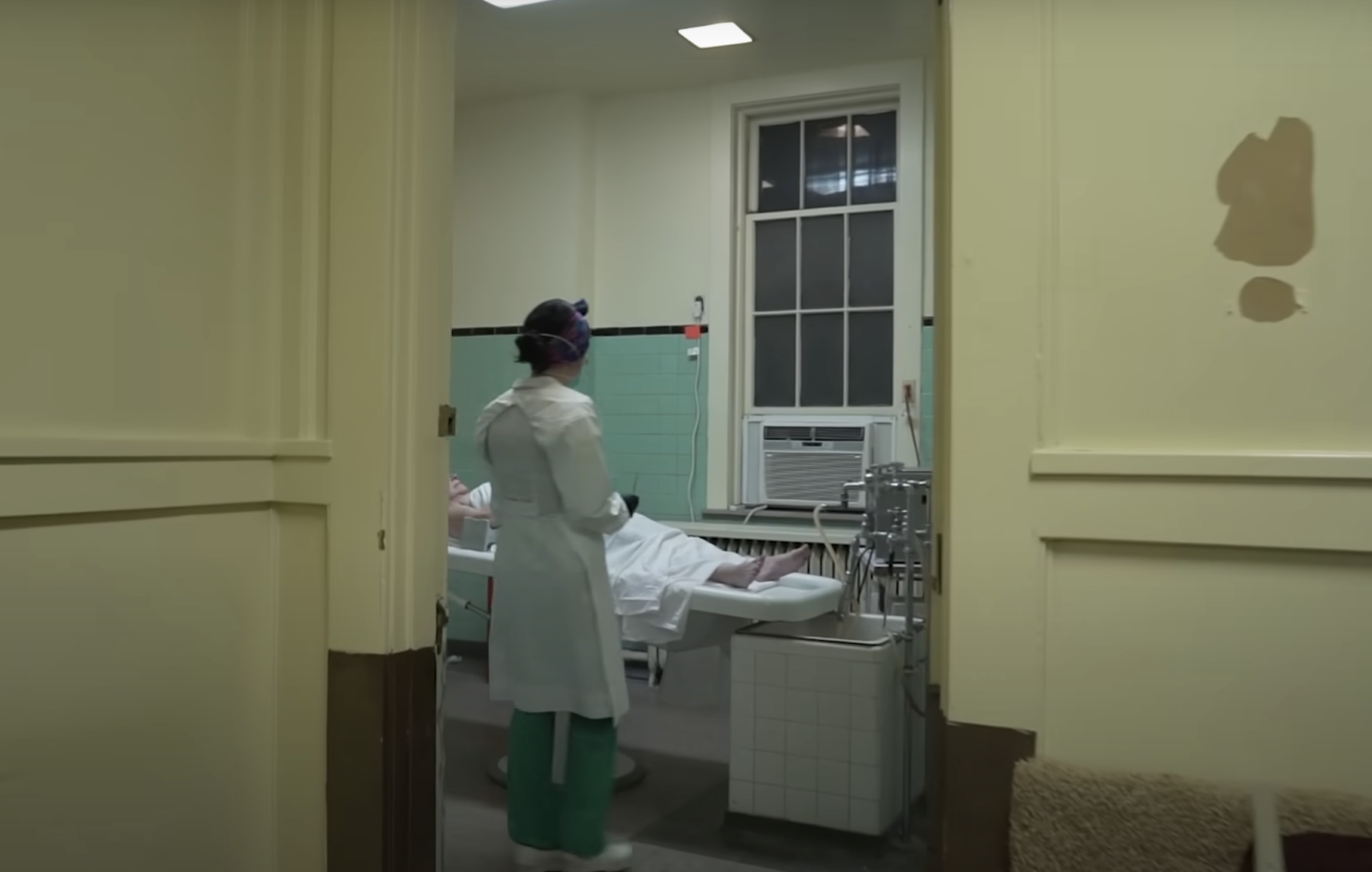 Person in medical attire stands next to a patient on a hospital bed in a room with medical equipment