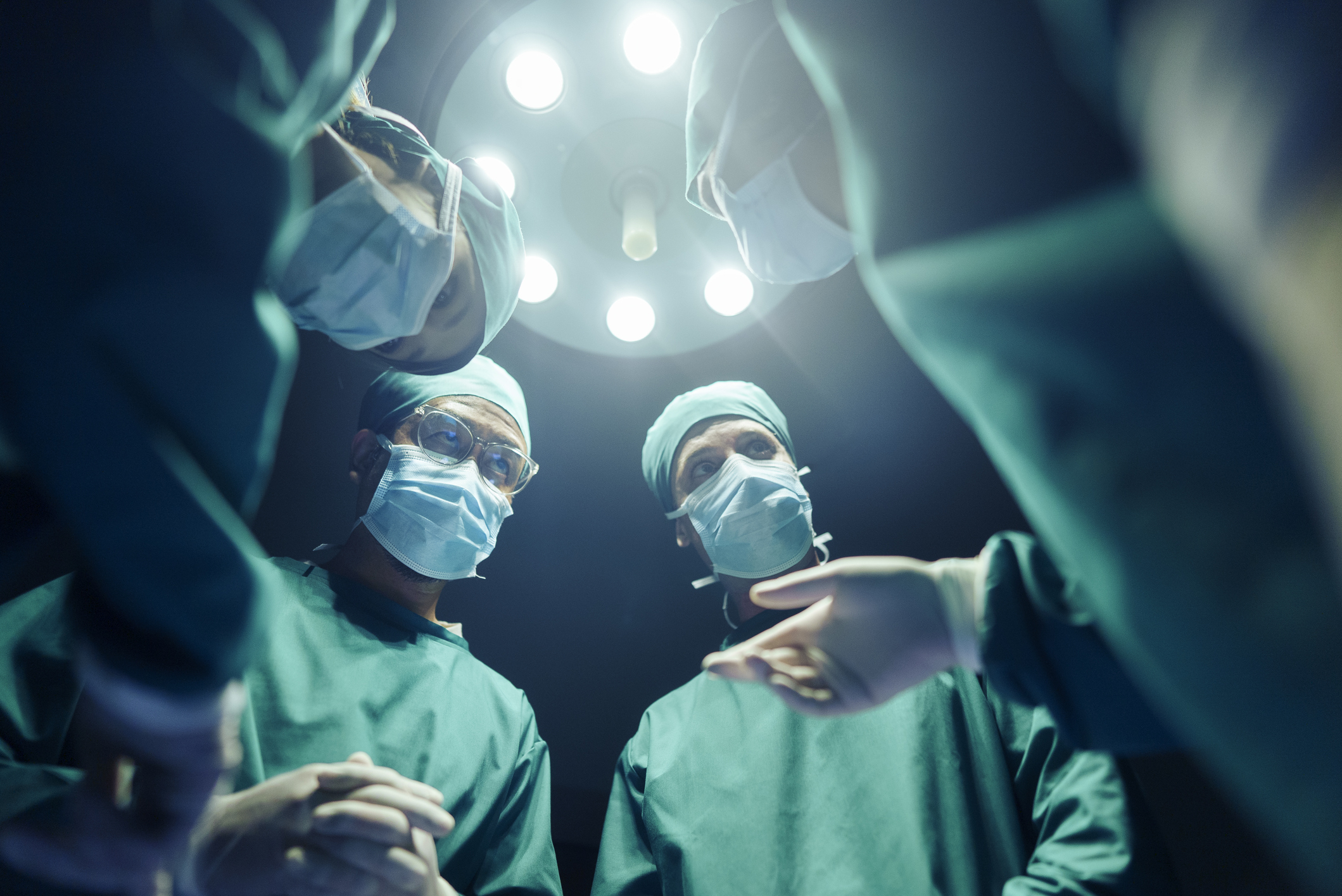 Surgeons wearing blue scrubs, masks, and gloves gathered under an operating room light, seemingly in the middle of a surgery or medical procedure