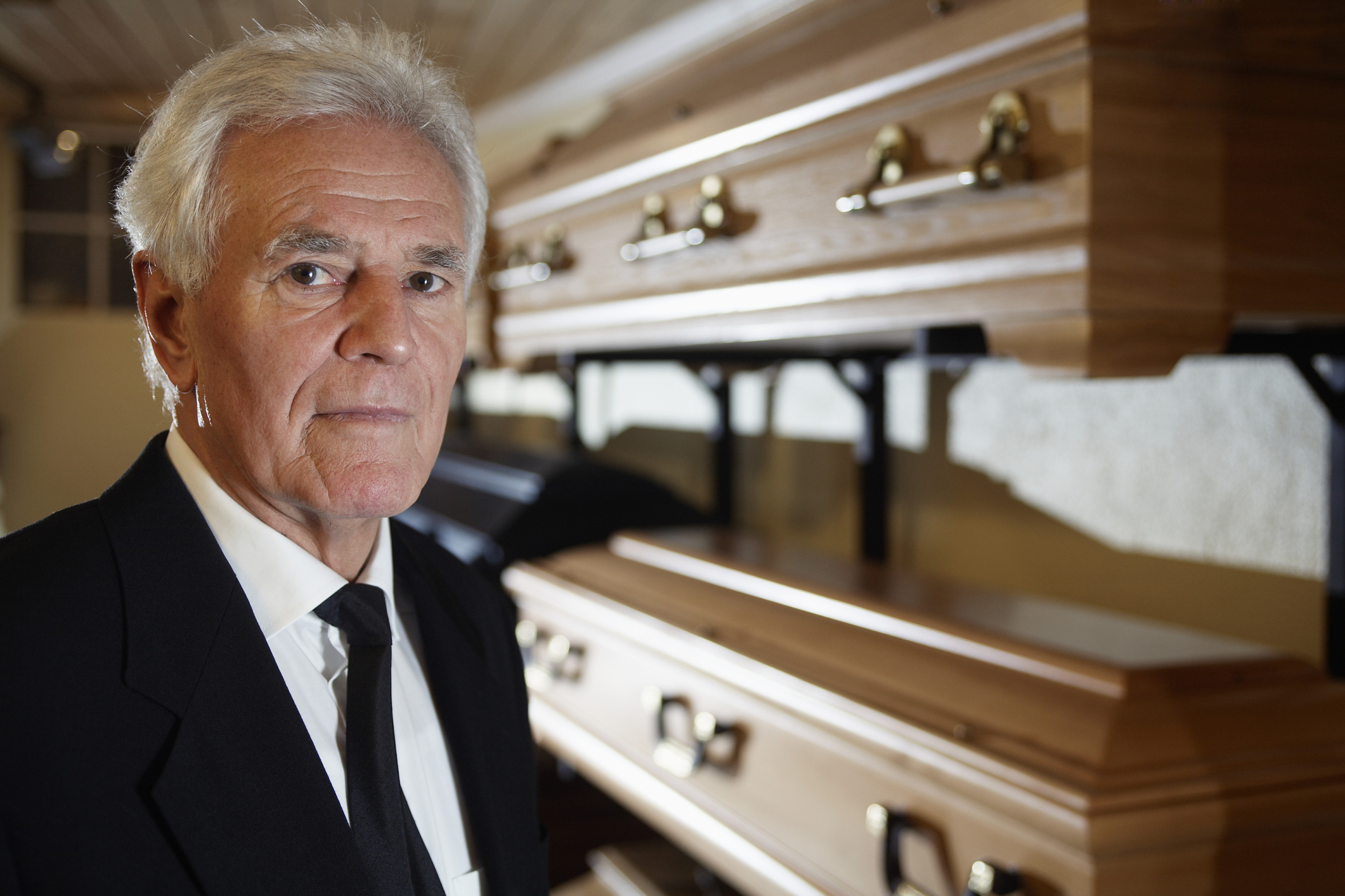 Older man in a suit standing beside various caskets, looking somber. The image relates to parents and final arrangements
