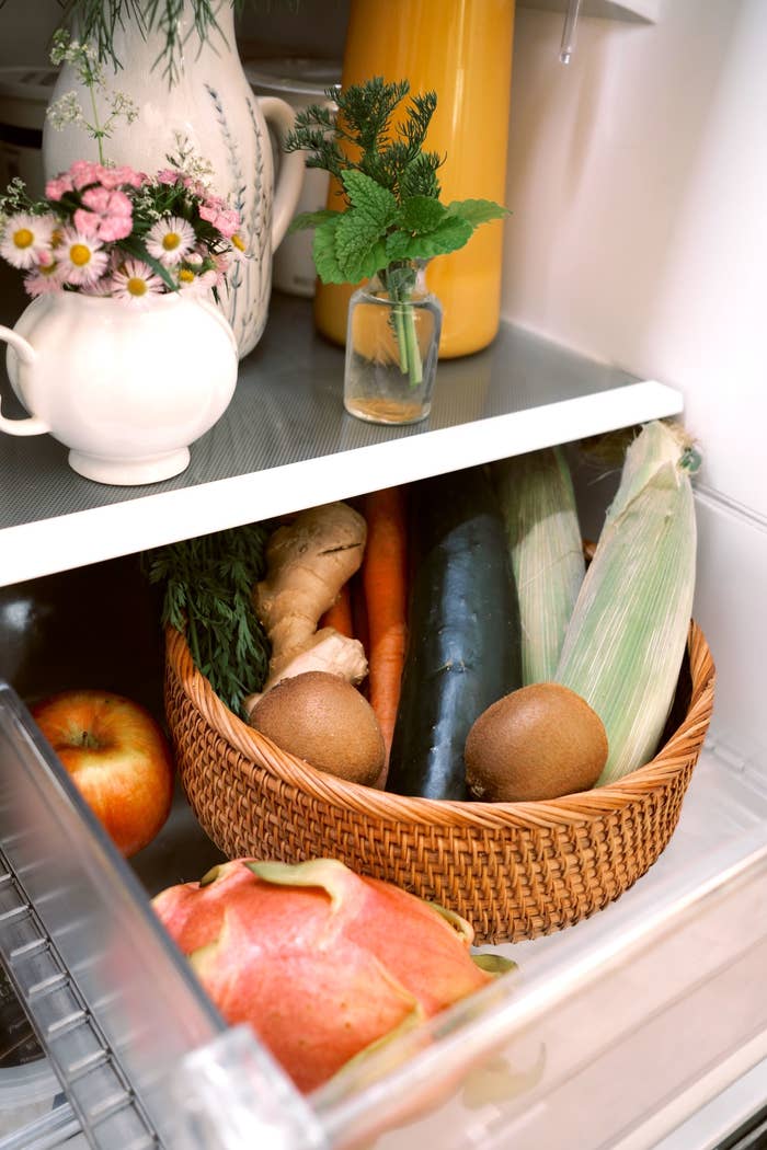 A refrigerator shelf with a basket containing ginger, carrots, zucchini, kiwis, corn, and an apple. There are also flowers and a yellow drink bottle