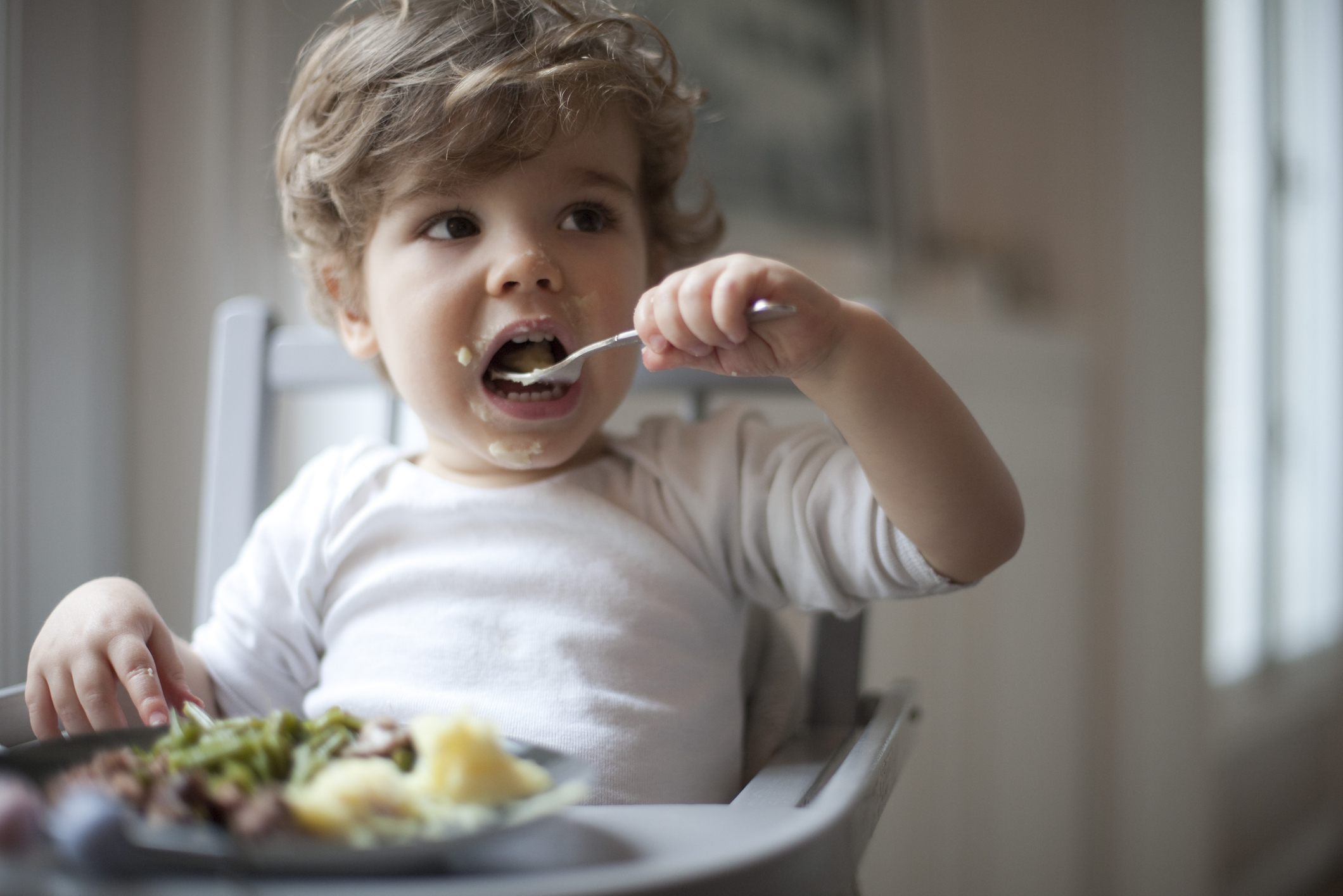 A small child with curly hair, wearing a white shirt, sits in a high chair while eating a meal using a spoon