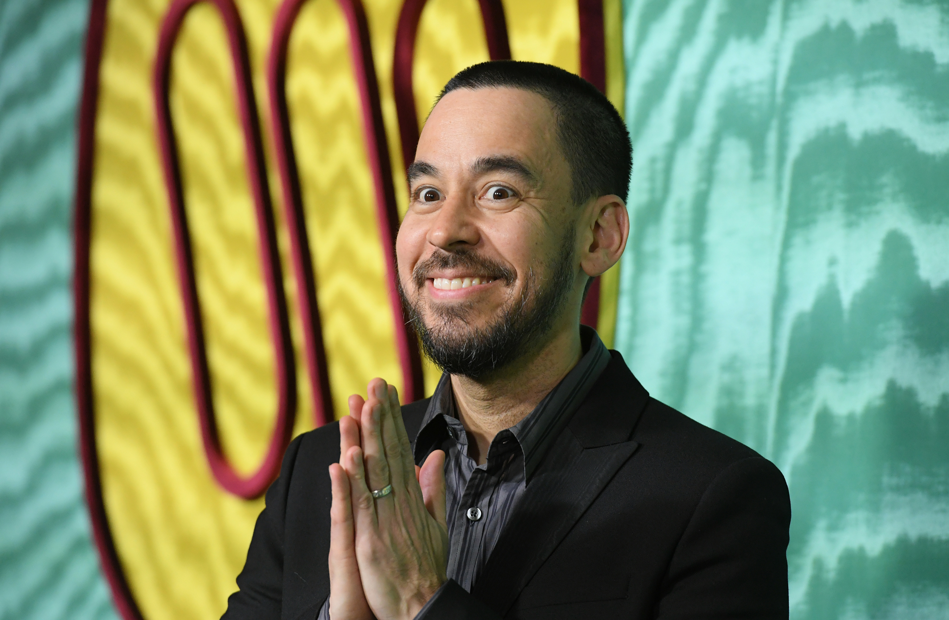 Mike Shinoda smiling, hands together in prayer-like gesture, wearing a dark suit and dark shirt, standing in front of an abstract background