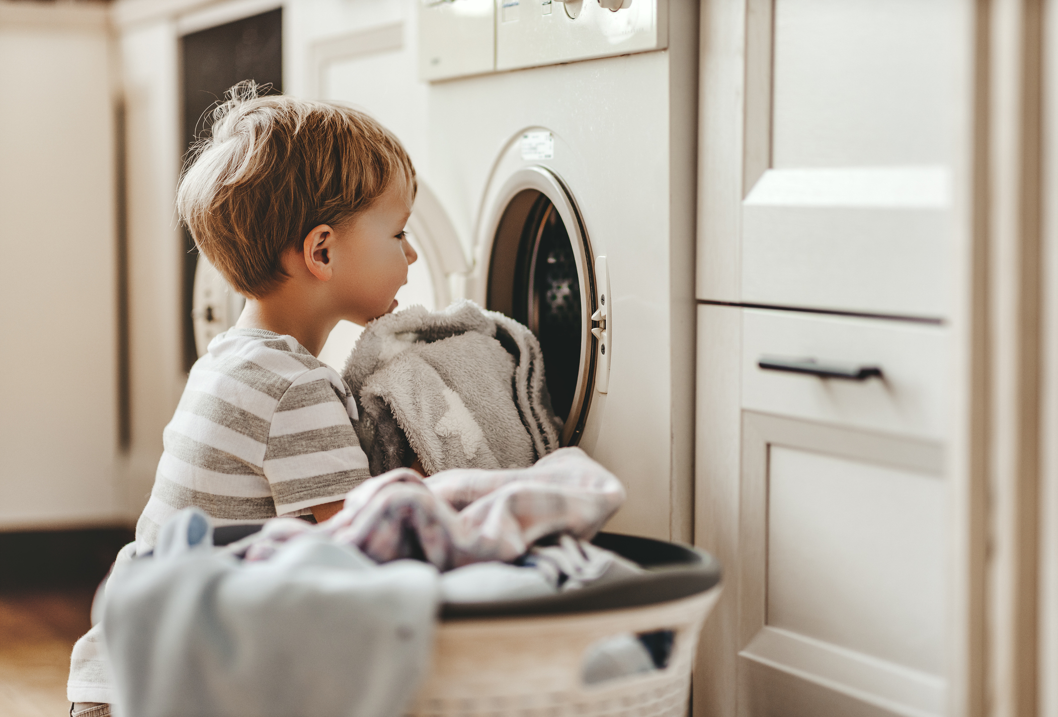 A young child wearing a striped shirt stands in front of a washing machine, putting clothes from a laundry basket into it