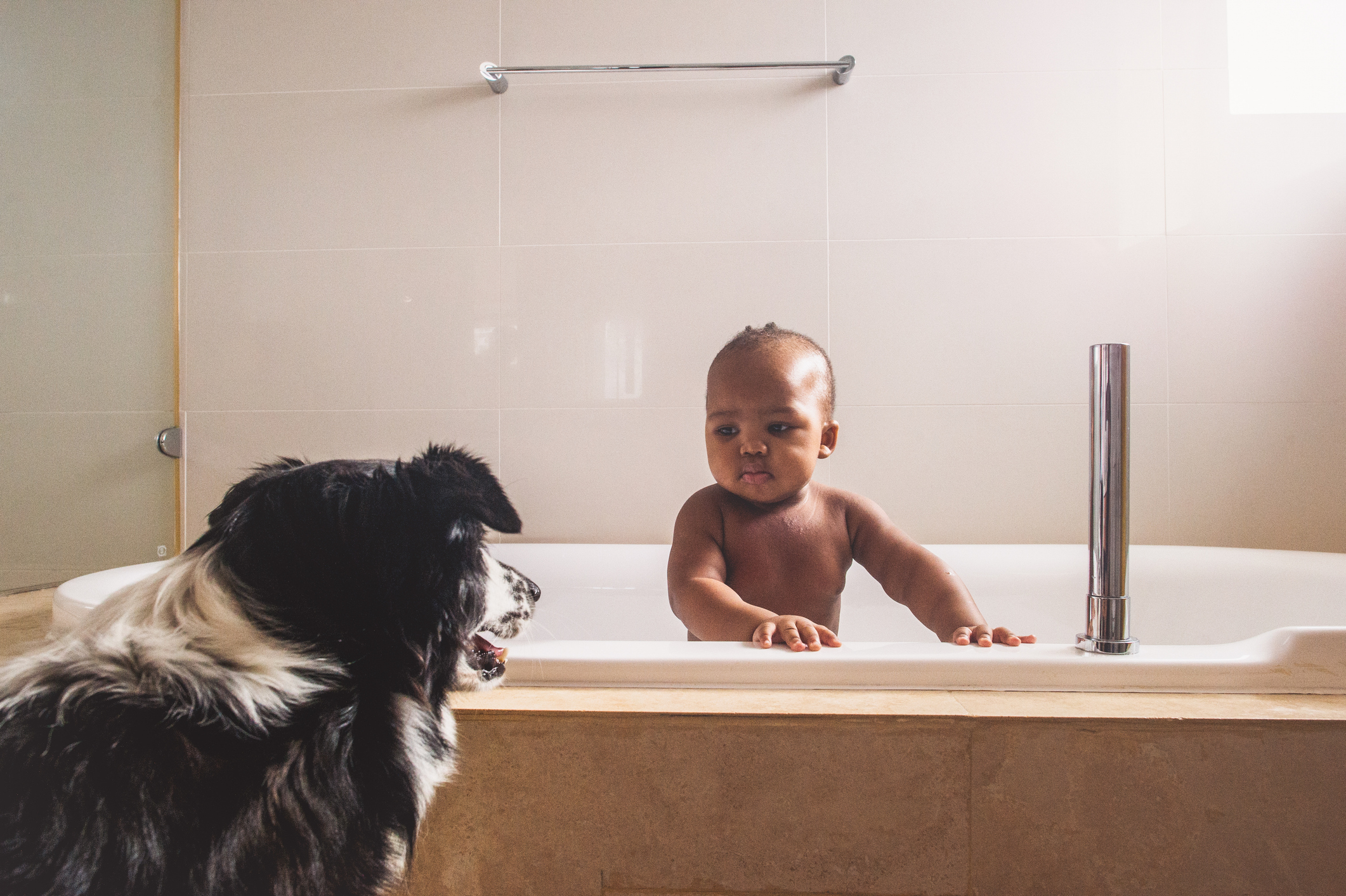 A baby bathing in a tub is looking at a dog standing next to the tub. The baby appears curious while the dog looks attentive