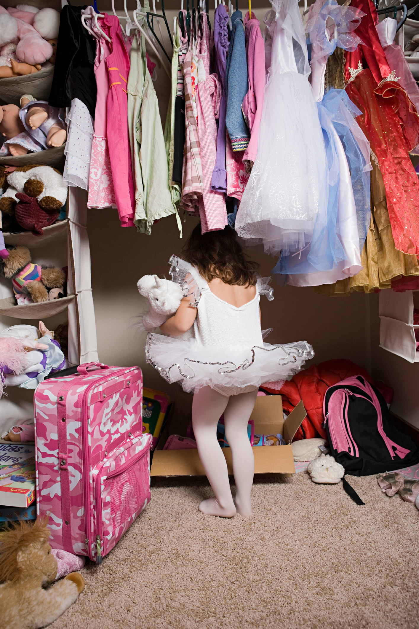 A young girl in a white tutu and tights stands in front of a closet filled with various clothes, holding a stuffed animal. A pink suitcase is on the floor
