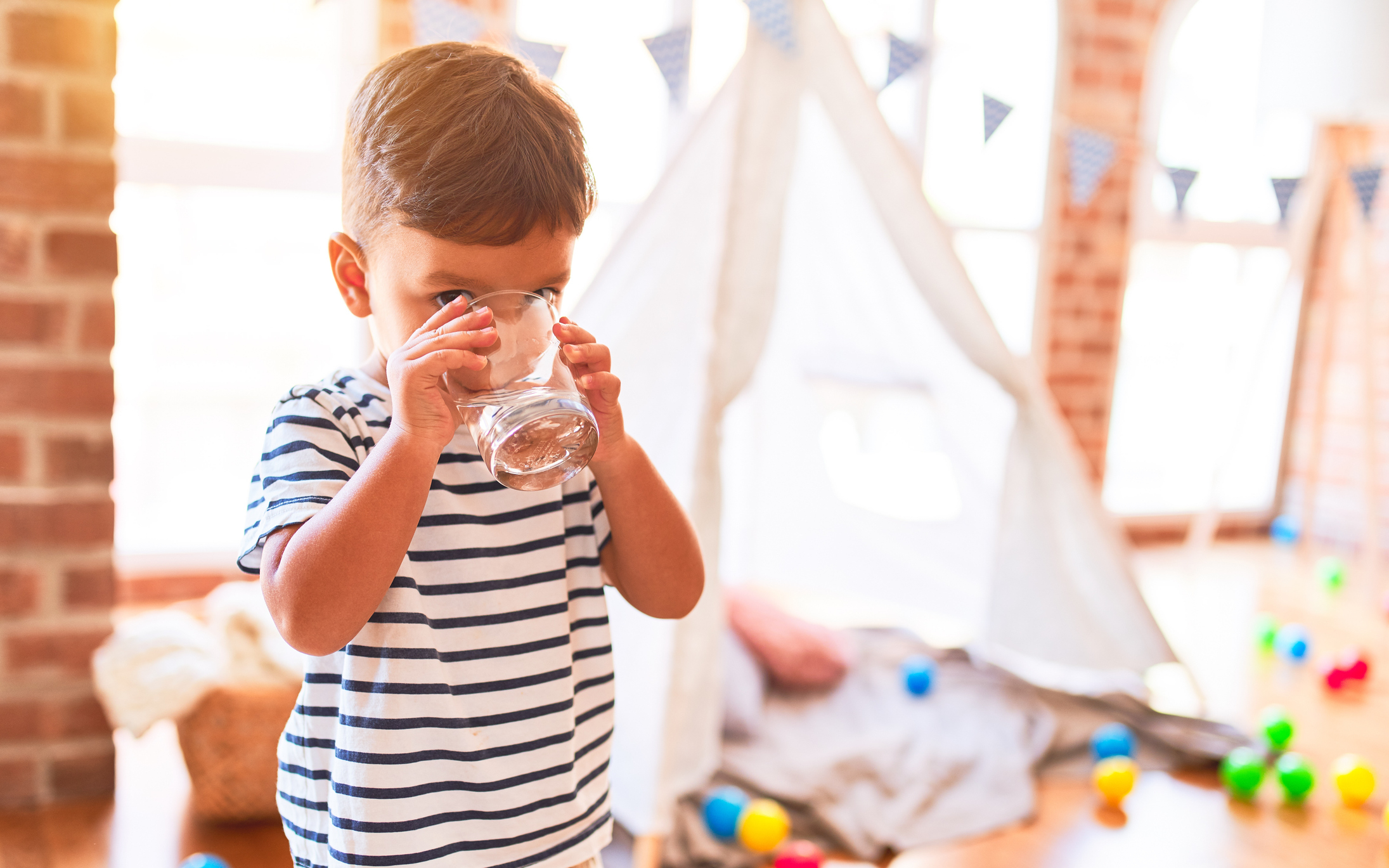A young child in a striped shirt drinks from a glass inside a room with a play tent and scattered toys in the background