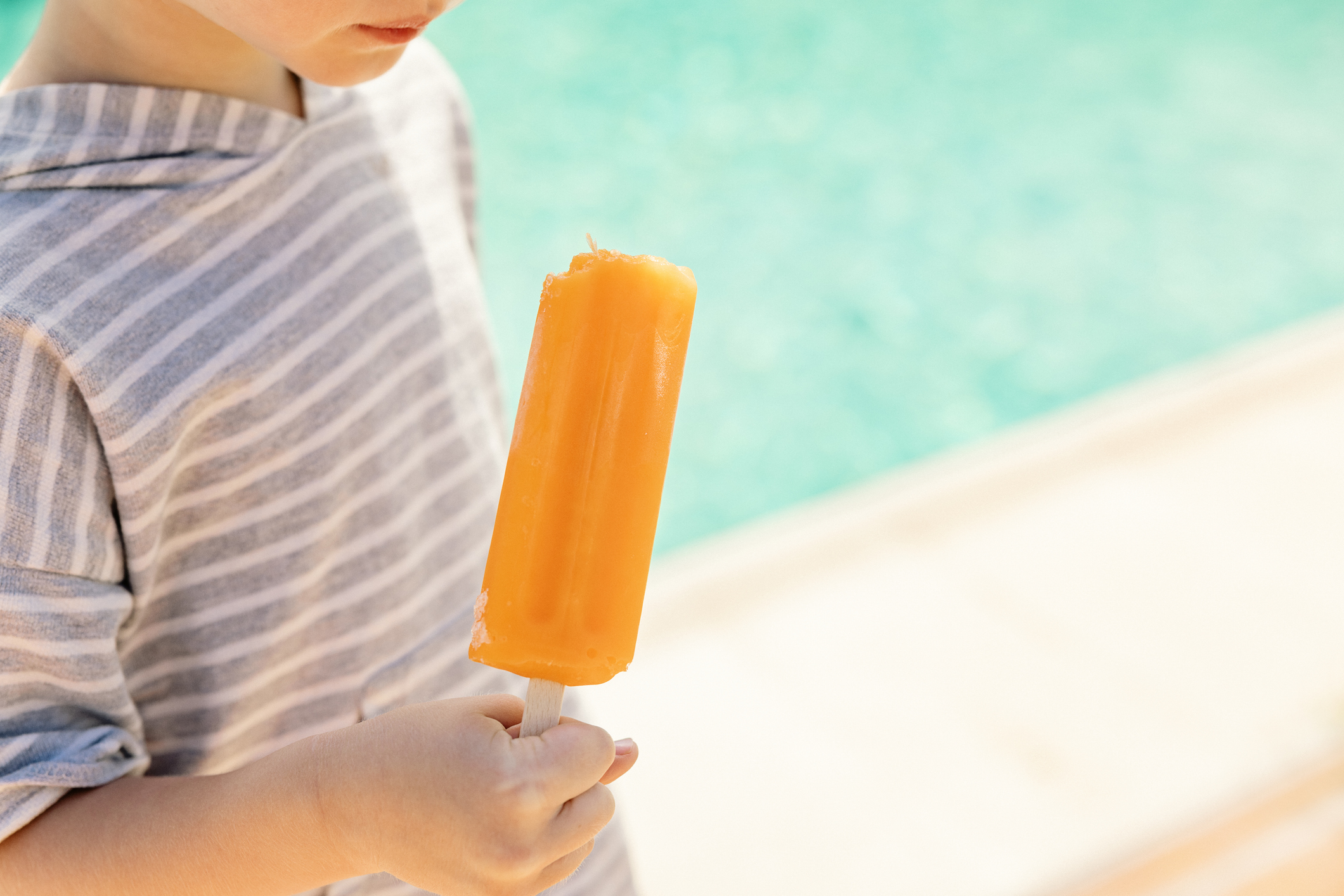 Young child in striped shirt holding and looking at a partially eaten orange popsicle by a pool