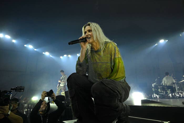 Emily Armstrong performs on stage, wearing a mesh top and cargo pants, holding a microphone, with band members playing instruments in the background