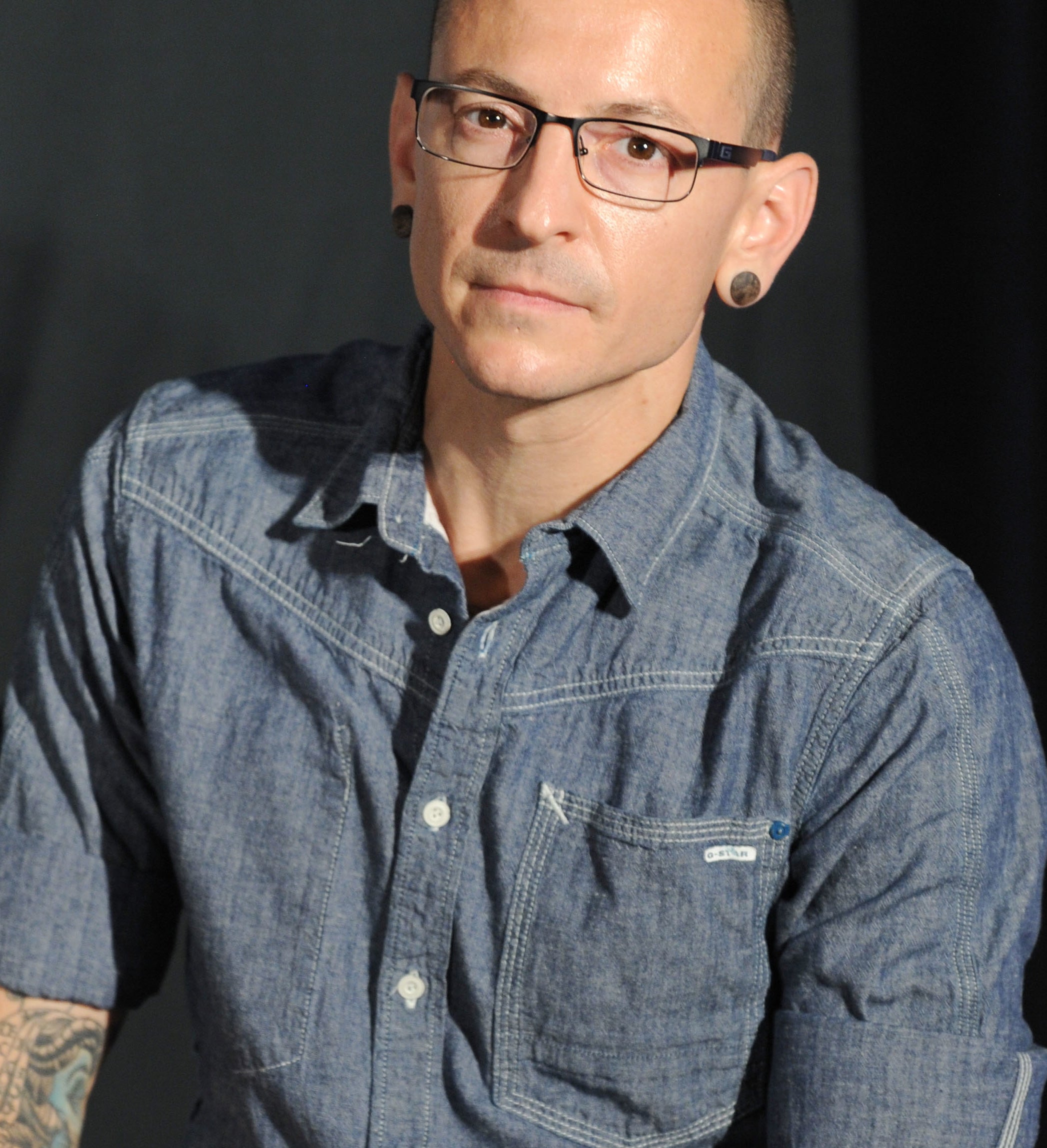 Chester Bennington with short hair and glasses, wearing a rolled-up denim shirt and showing arm tattoos, stands against a plain background